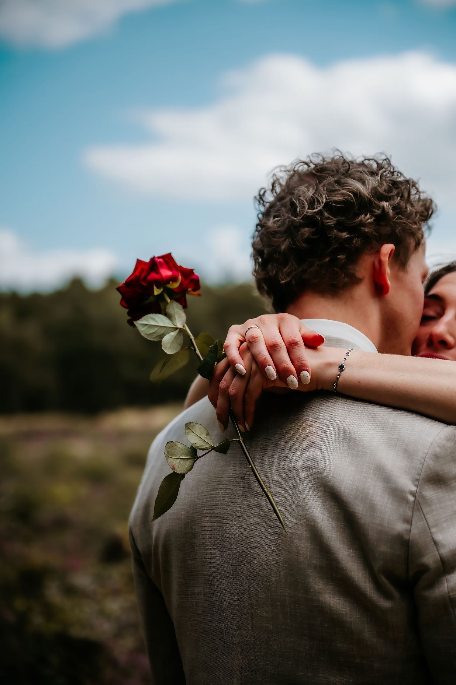 A single red rose held during an intimate embrace against a blue sky backdrop.