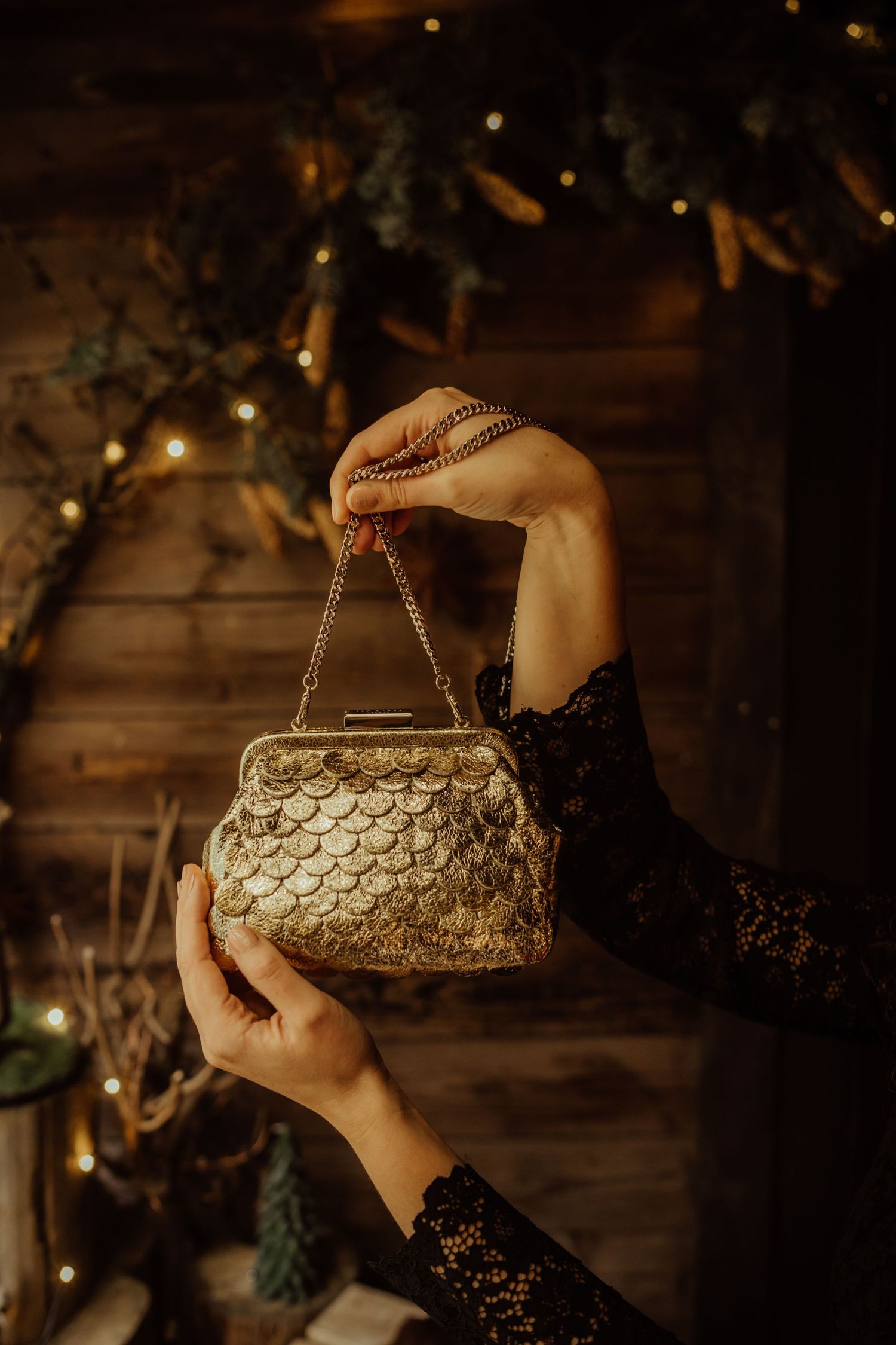 Hands in black lace sleeves hold a vintage gold sequined evening purse against a dark wooden wall with holiday lights.