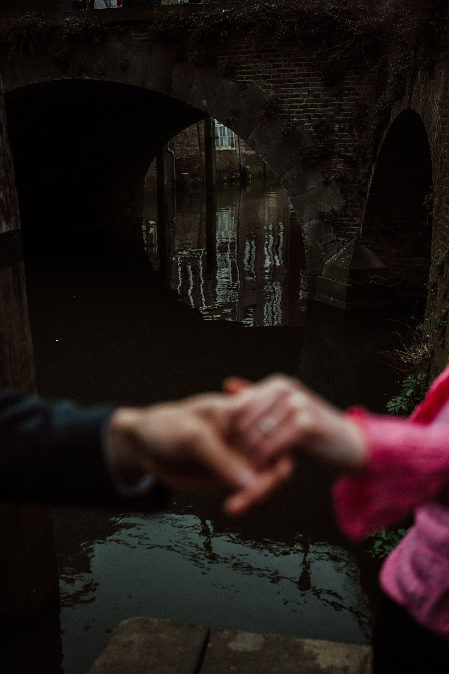 Two hands clasped together in front of a dark stone archway bridge with reflections in the water below.