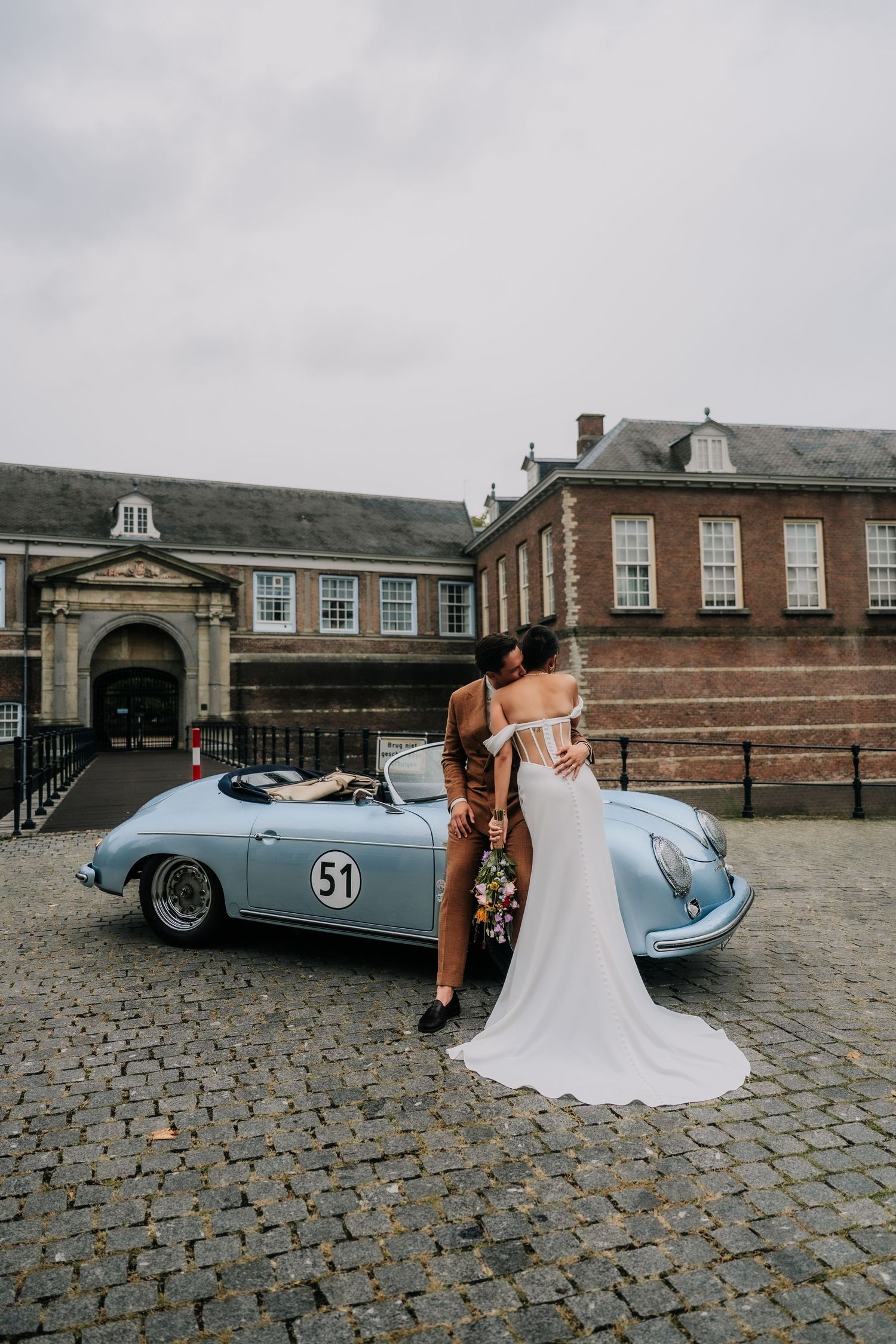 Couple embracing beside a vintage light blue Porsche 356 convertible on cobblestones outside a brick building.