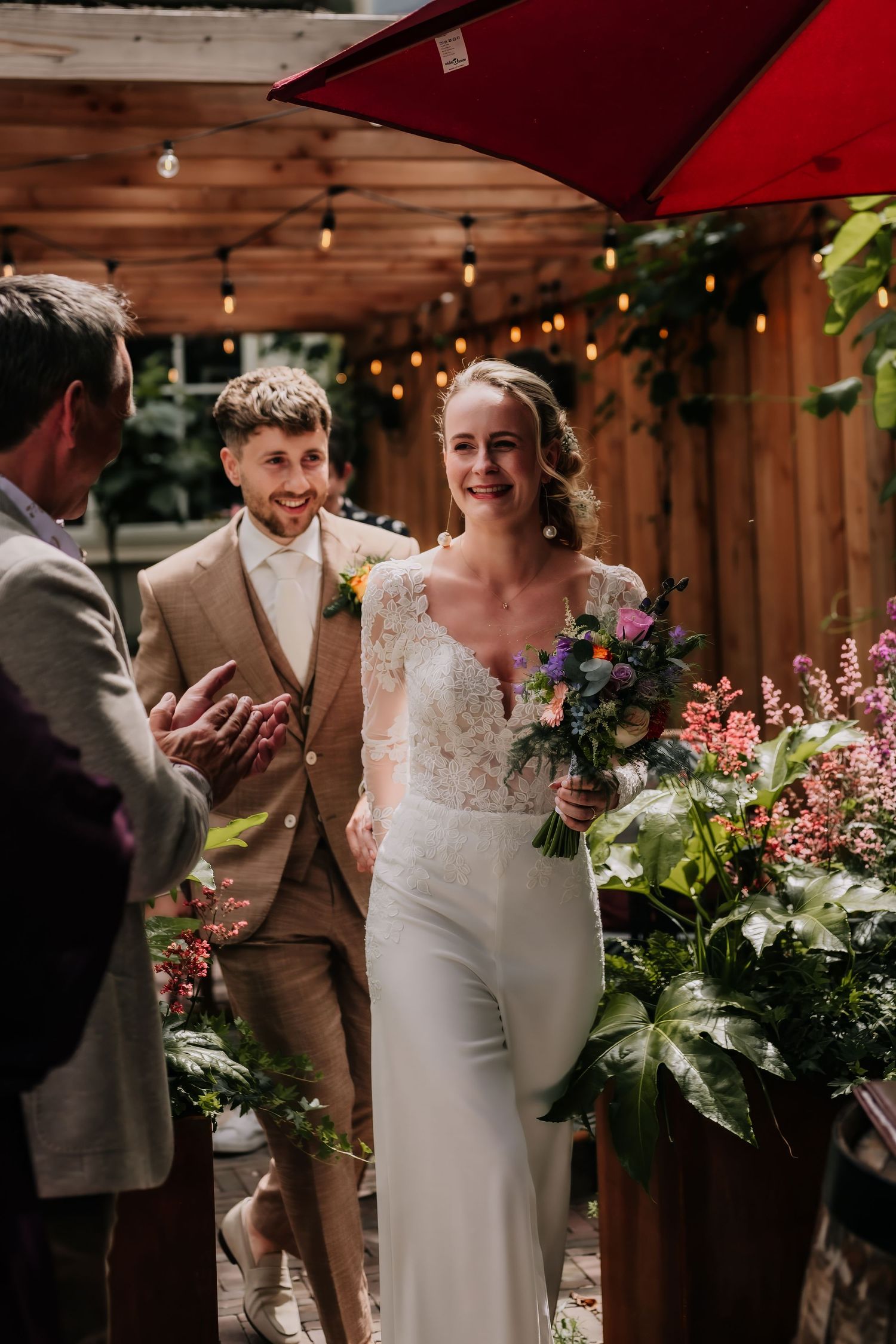 Smiling bride in white lace jumpsuit holding colorful bouquet walks with groom in tan suit at outdoor wedding ceremony.