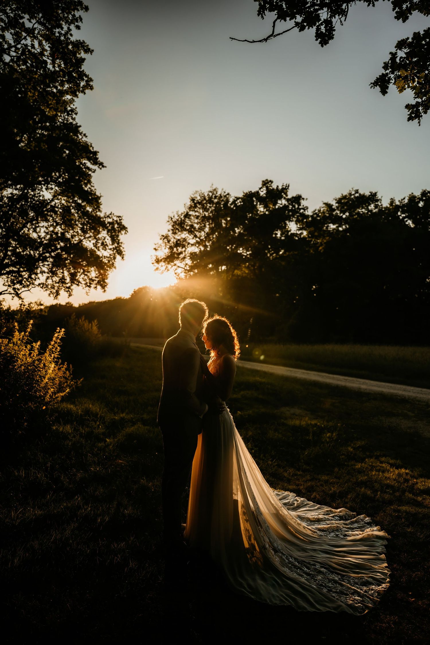 Silhouetted figures in wedding attire stand against a dramatic golden sunset with flowing dress train creating ethereal glow.