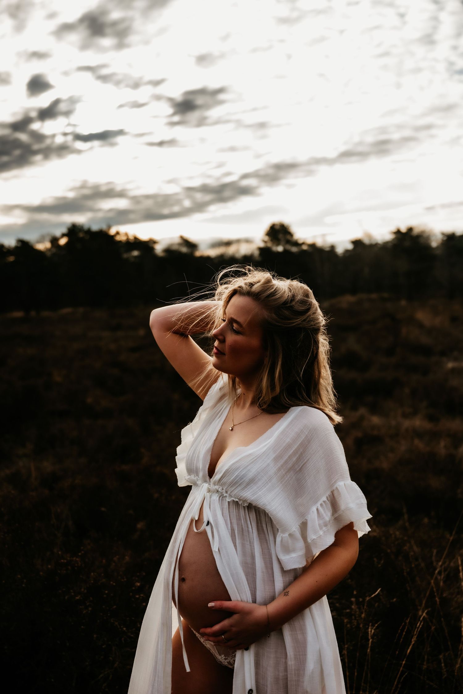 A person in a white flowy dress poses in a field at sunset cradling a pregnant belly thoughtfully. Mastbos, Brda, zwangerschapsshoot.