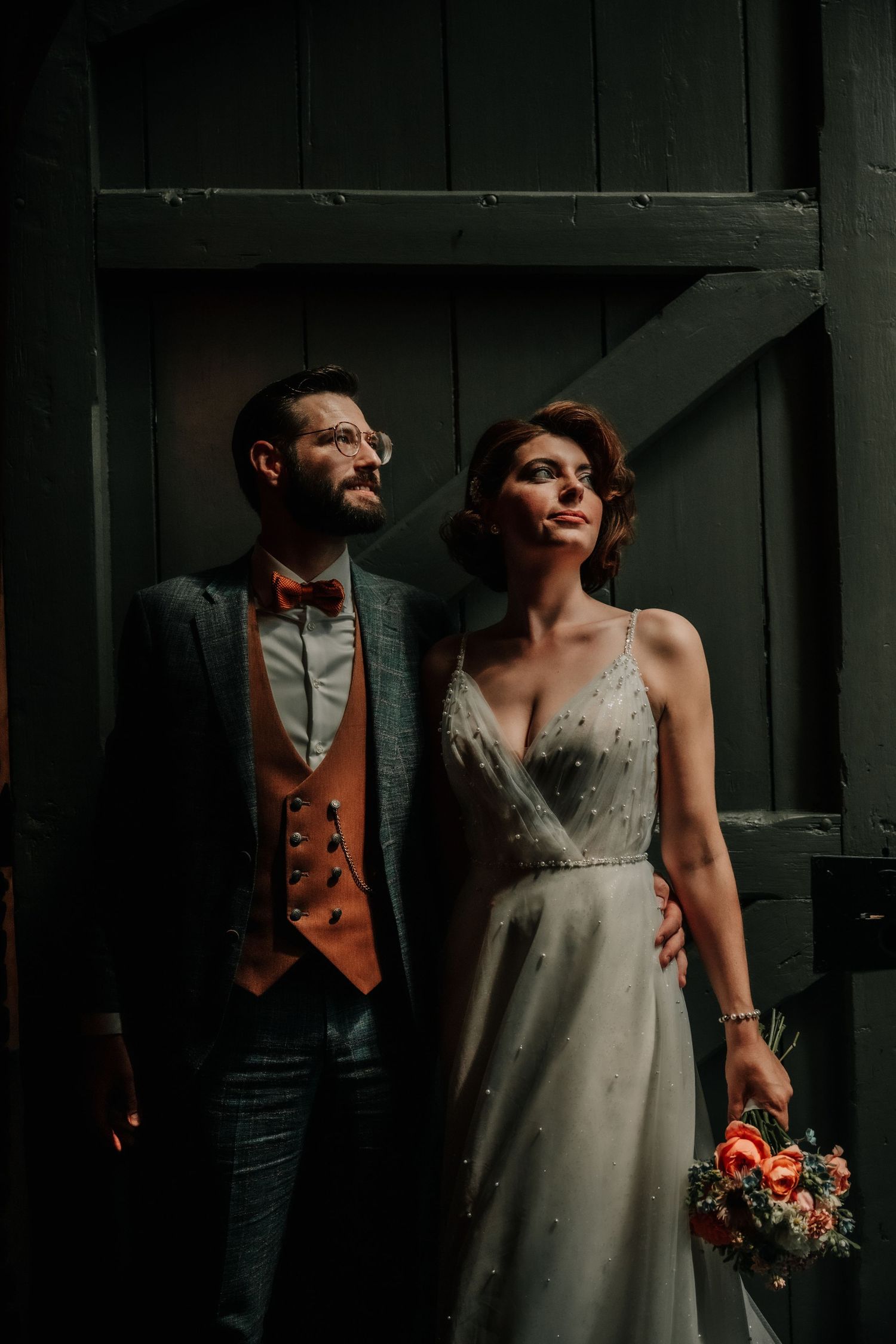 Couple in formal wedding attire share intimate moments in a moody indoor setting with dramatic lighting.