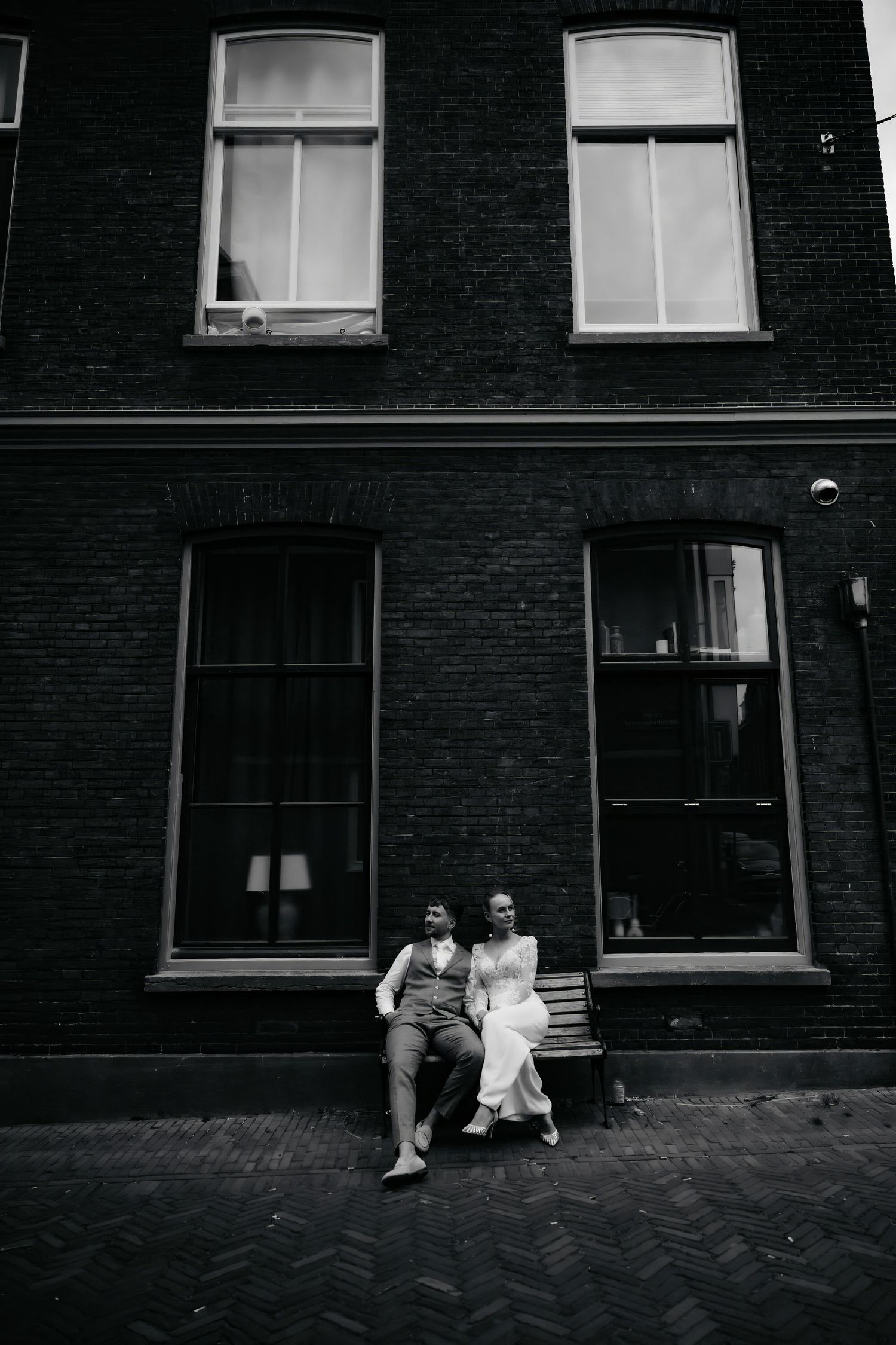 Black and white photo of a bride and groom sitting on a bench outside a brick building.