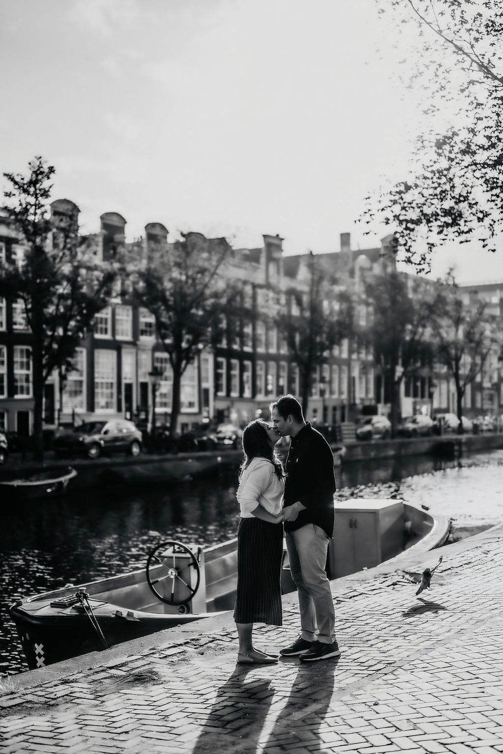 Couple embracing by Amsterdam canal with boats and historic buildings in black and white photo.