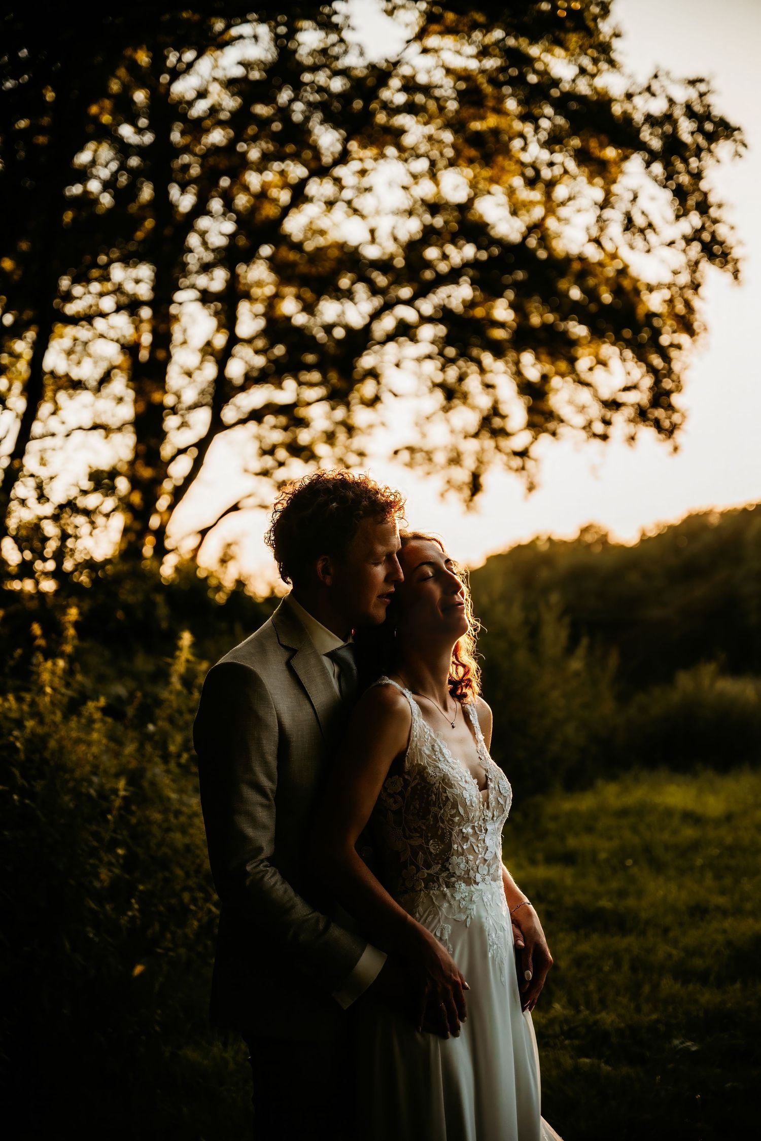 A couple shares an intimate moment during golden hour sunset in a field with trees silhouetted in the background.