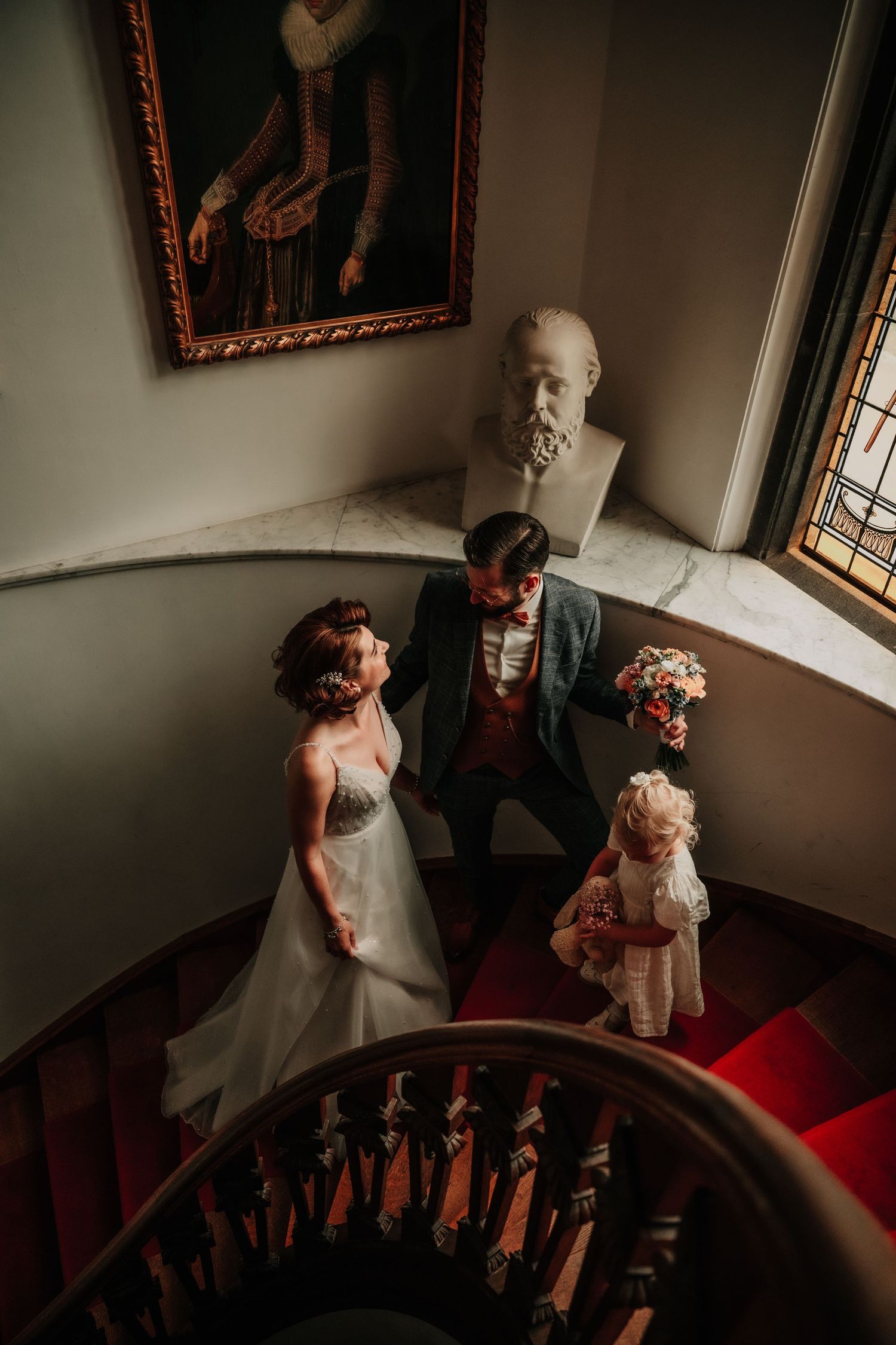 A wedding couple poses on elegant red-carpeted stairs with historic portraits and a marble bust in the background.