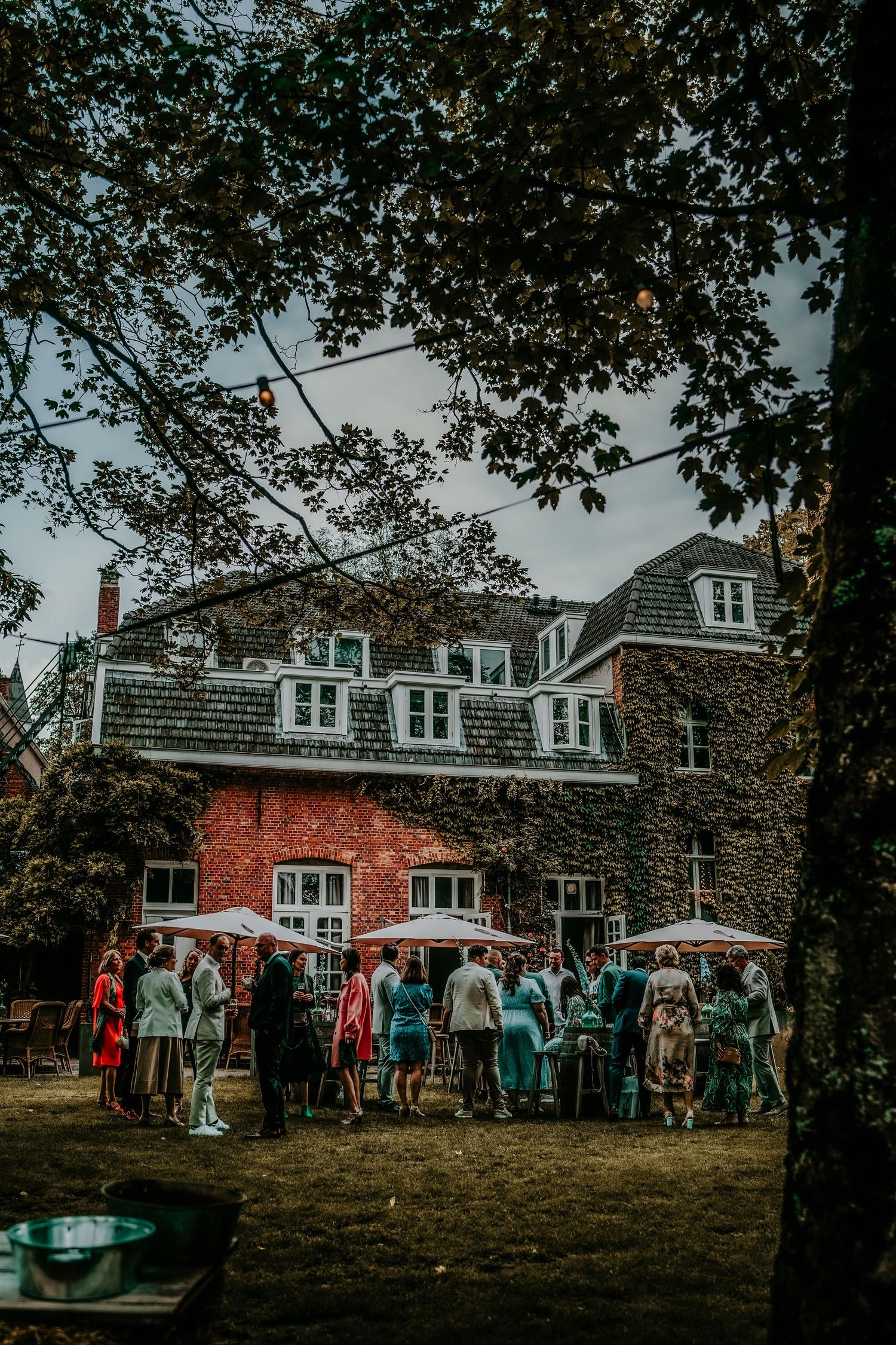 A group of people gathered outside a red brick house with dormers during an outdoor event on a sunny day.