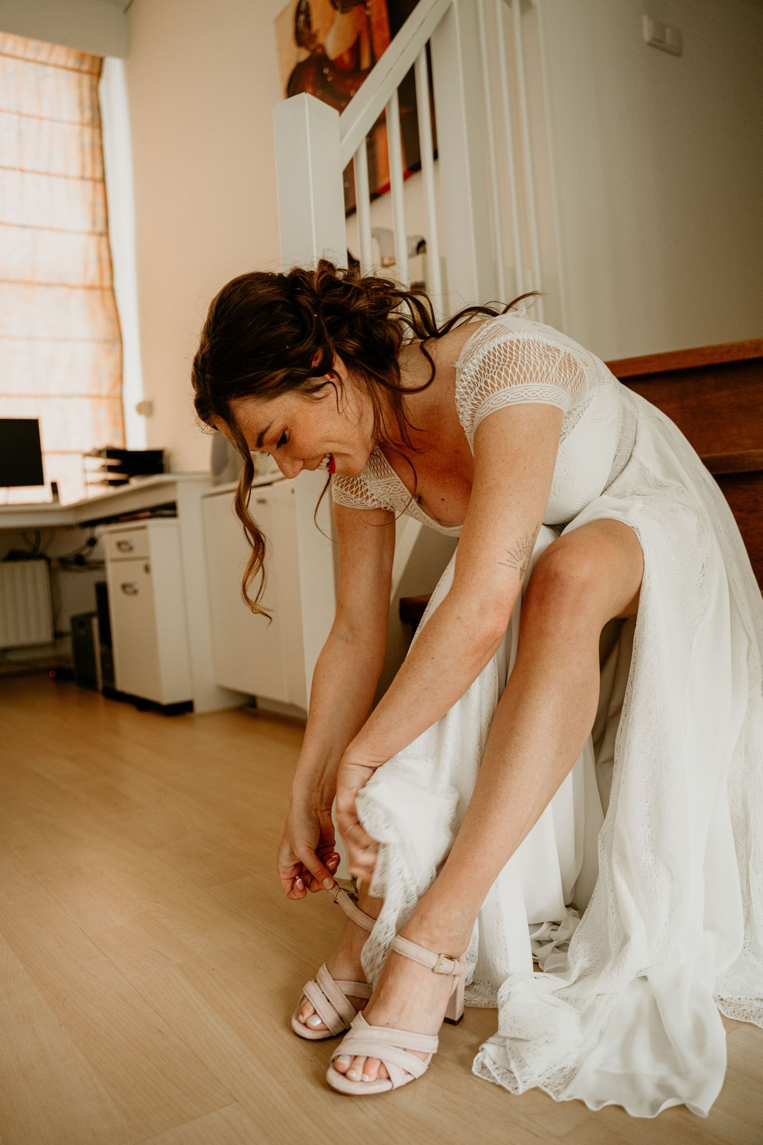 Bride adjusting elegant white sandals while seated in a bright room.