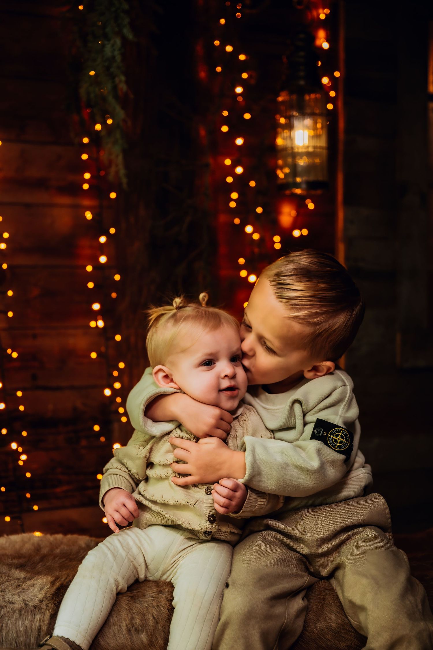 Two young children in matching outfits share tender moment on wooden bench with holiday lights in background.