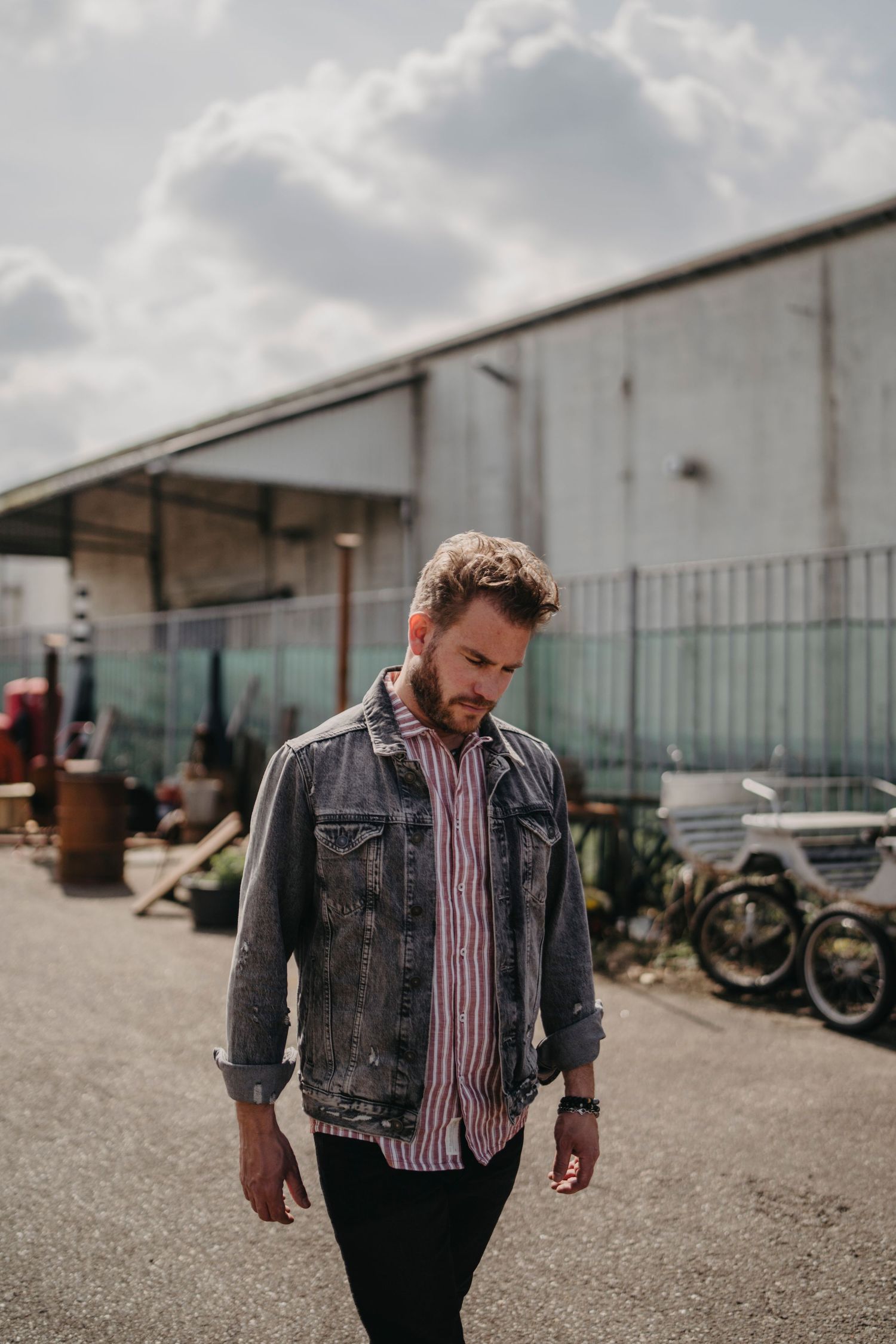 A person in a denim jacket walks along a gravel path next to industrial buildings on a cloudy day.