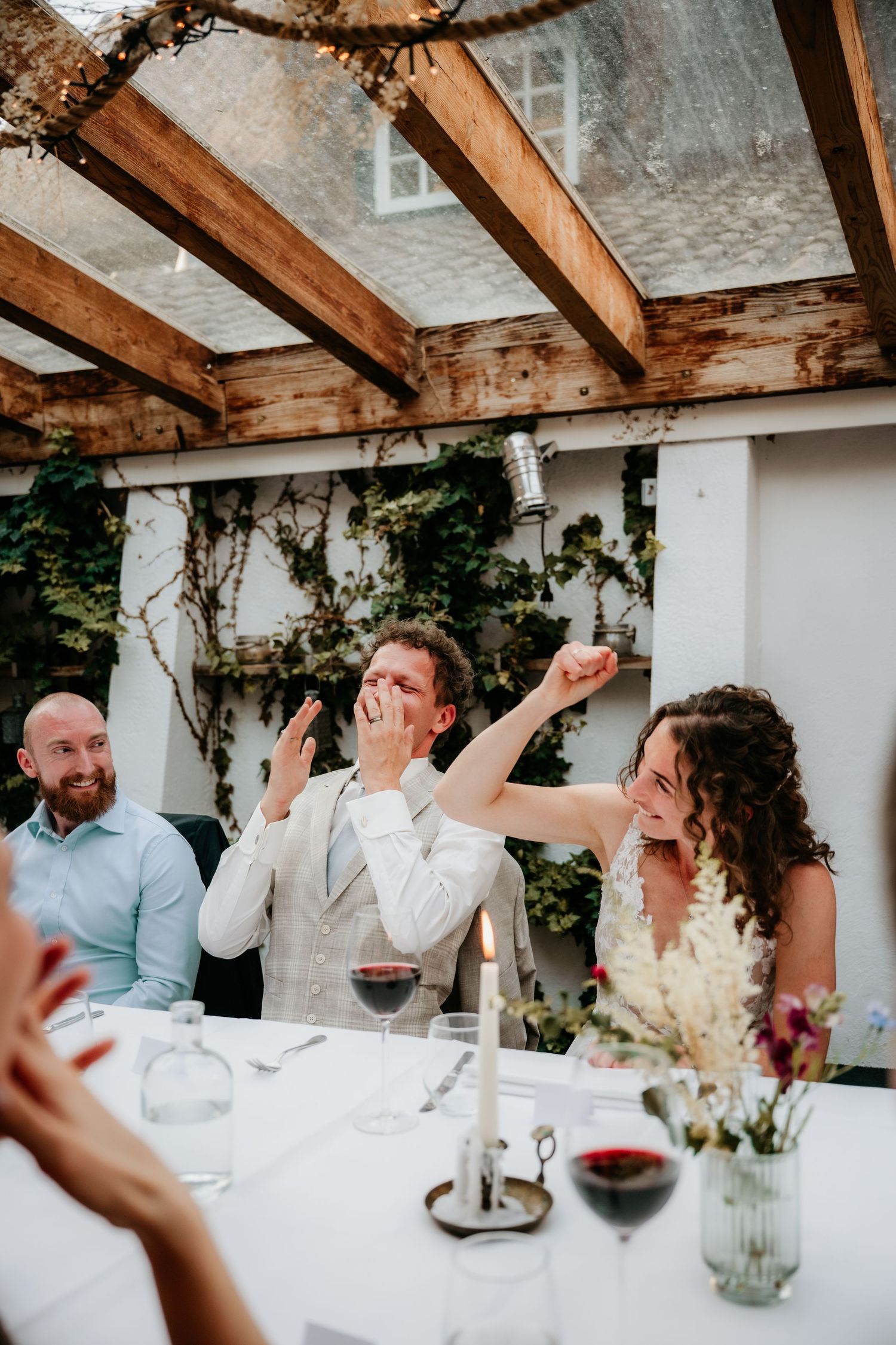 Wedding reception dinner with guests laughing and celebrating under wooden pergola.