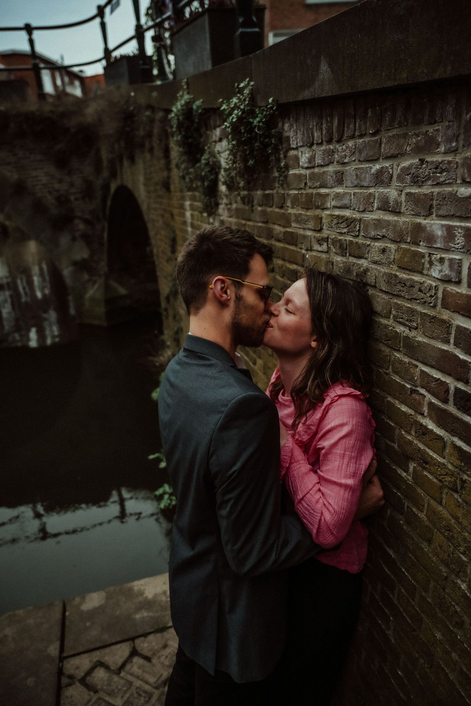 Romantic couple embraces against rustic brick wall in atmospheric low light setting.