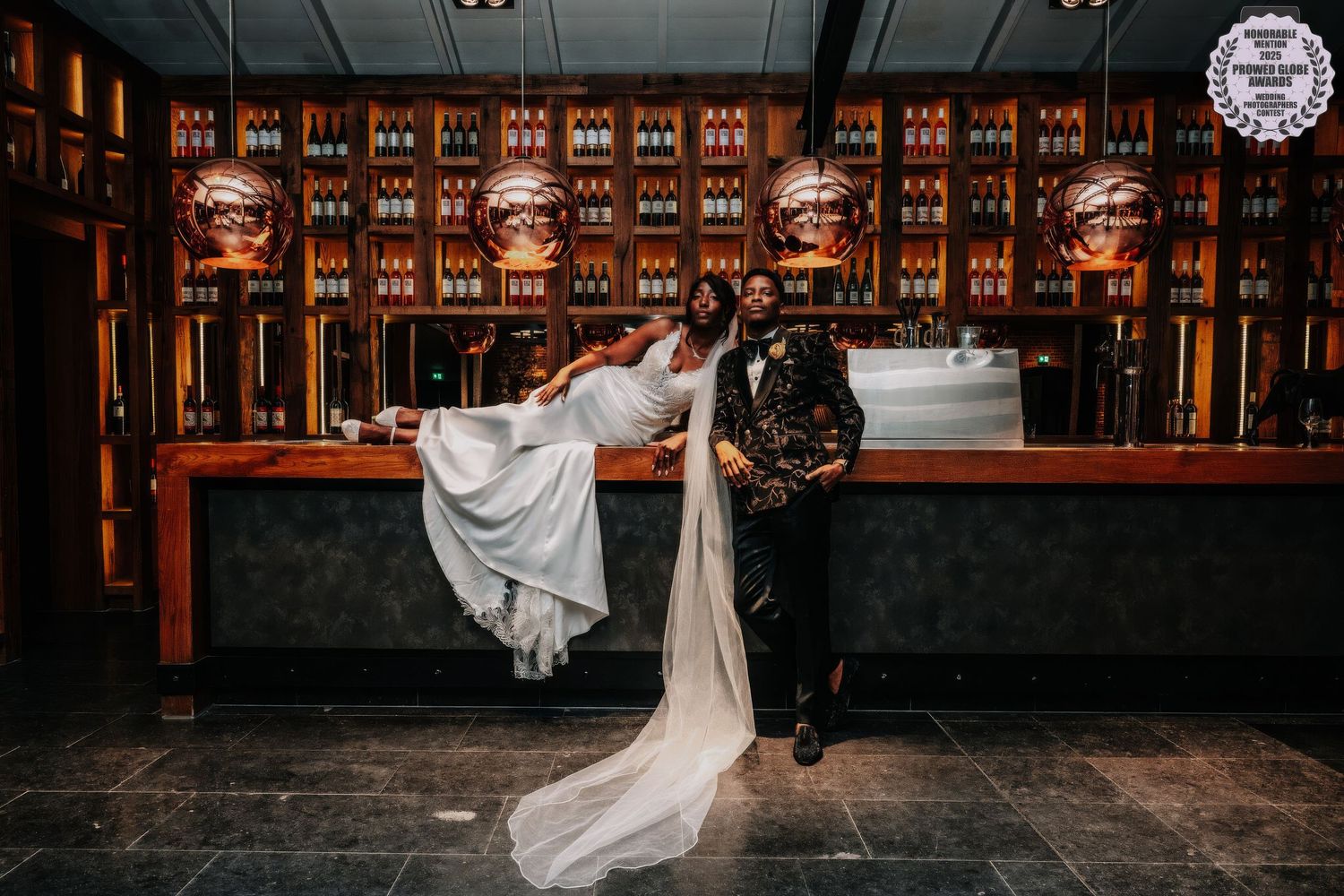 A couple in wedding attire dances together in front of a warmly lit rustic wooden barn venue at sunset.