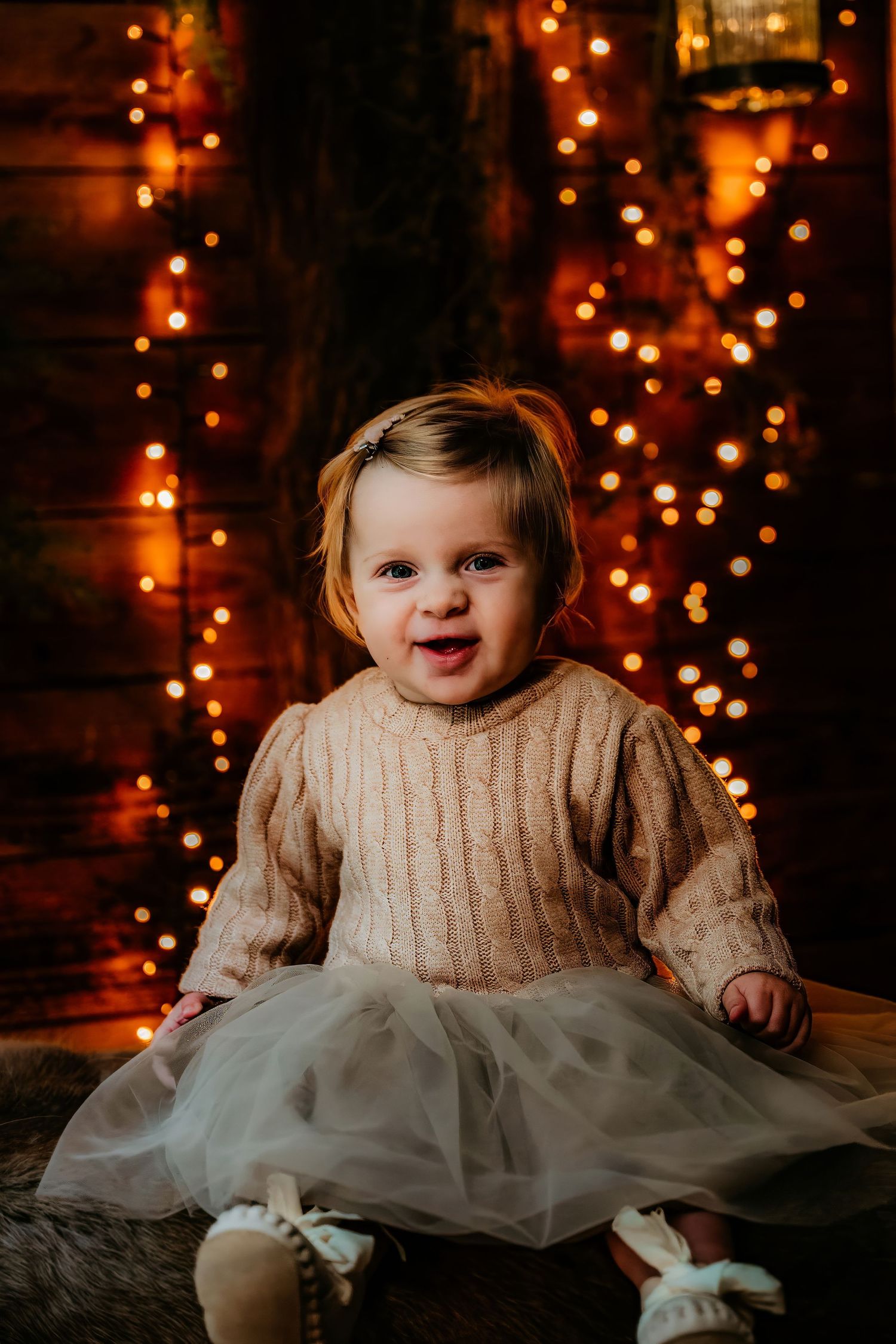 A small child in a cream sweater and tulle skirt sits against a backdrop of warm glowing Christmas lights.