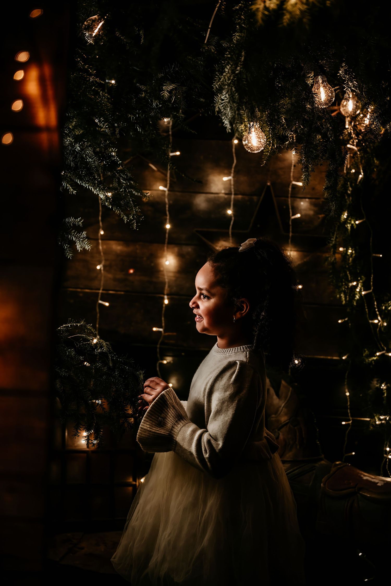 A silhouette stands in a dreamy setting with twinkling lights and evergreen branches creating a magical holiday atmosphere.
