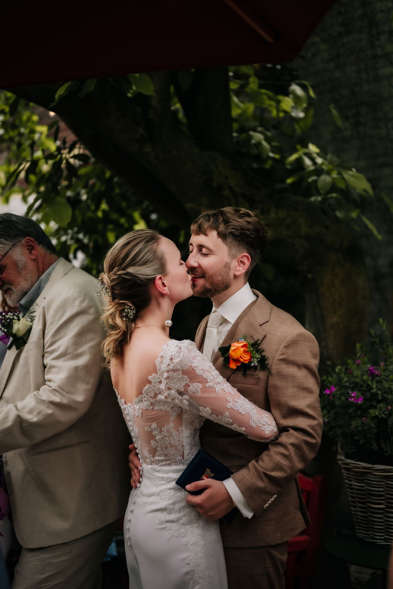 Bride and groom share an intimate moment at outdoor wedding ceremony, bride in lace dress, groom in tan suit with floral boutonniere.