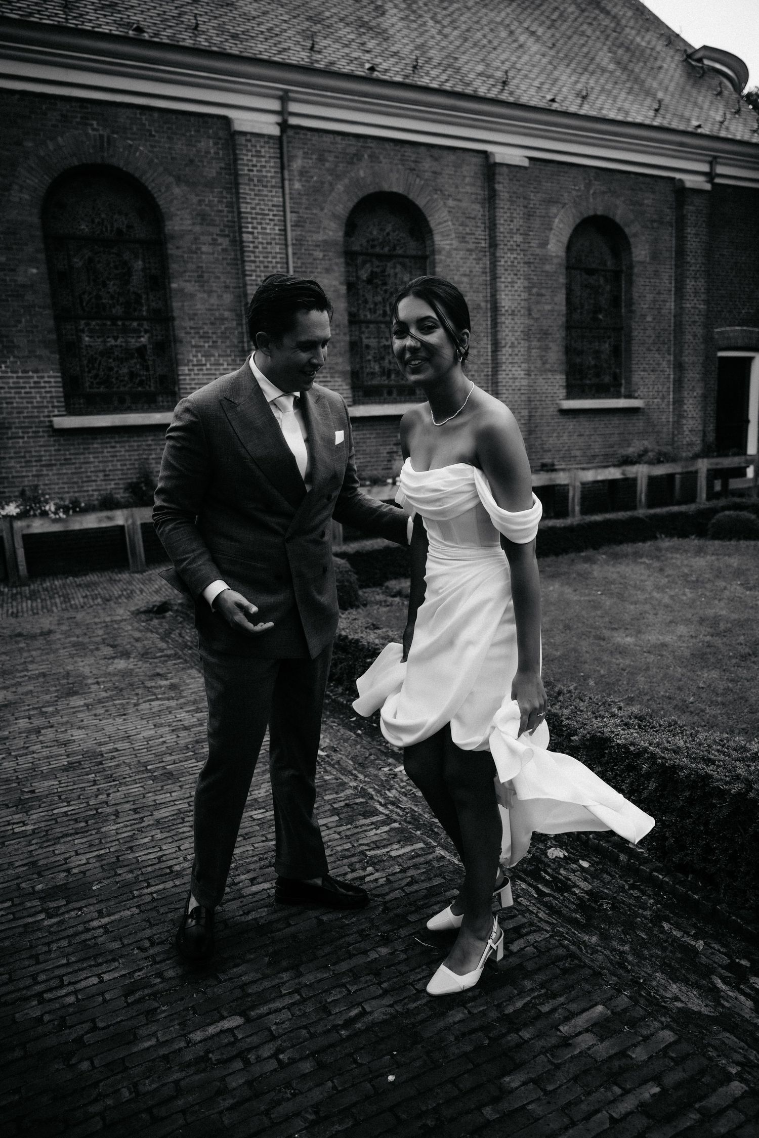 Black and white photo of a smiling bride and groom walking together outside a brick church on their wedding day.