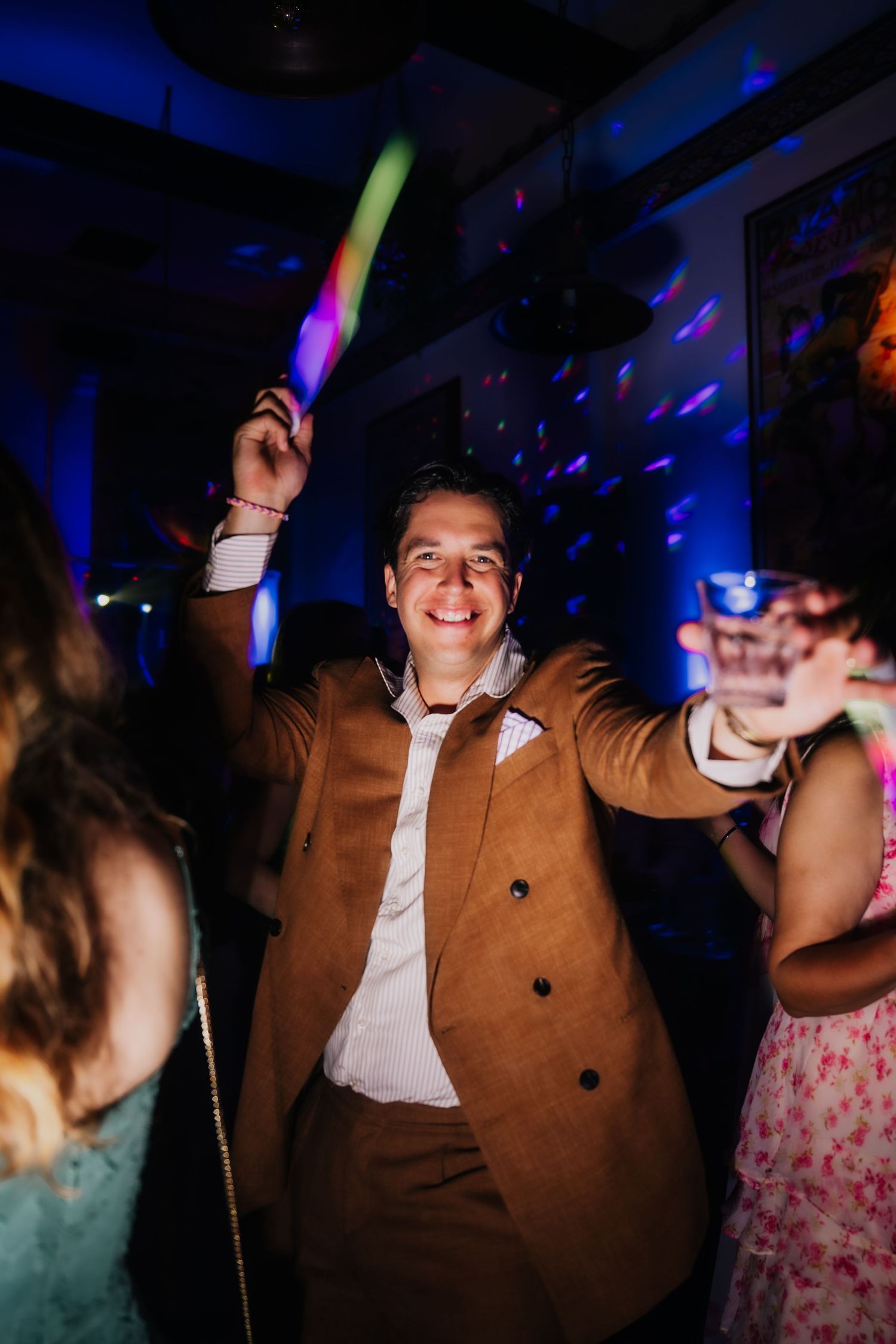 Man in brown suit dancing at party, holding glow stick under colorful disco lights.