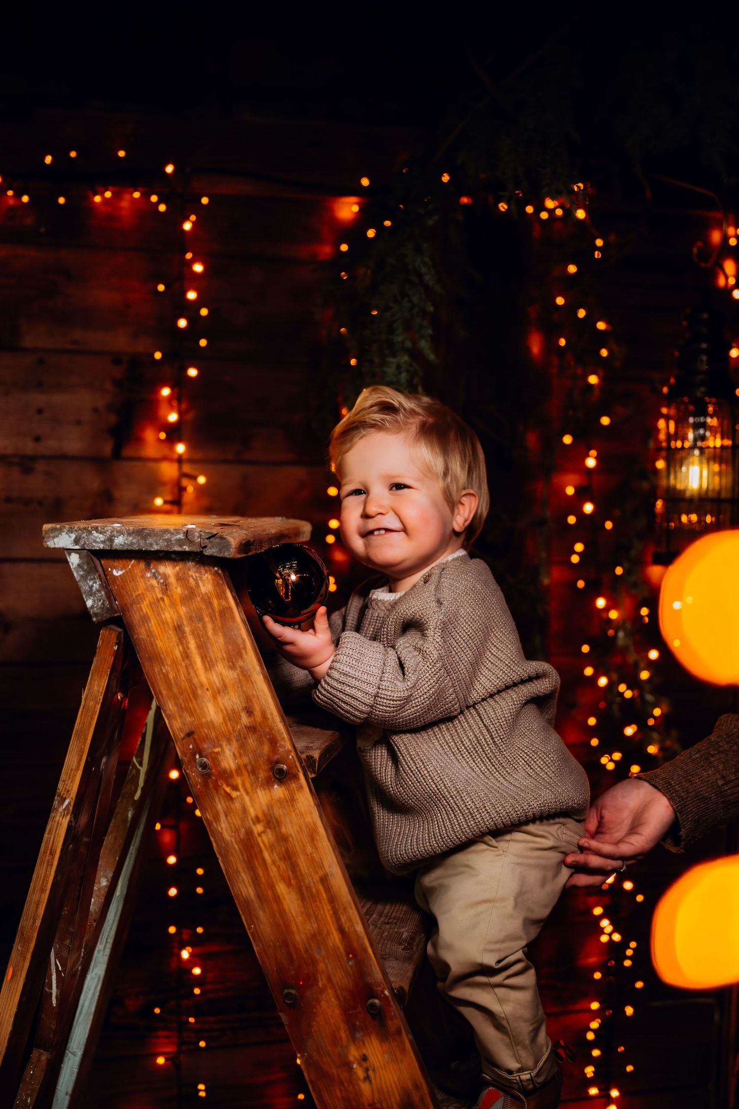 Young child in a sweater plays on a wooden chair against a backdrop of warm holiday lights.