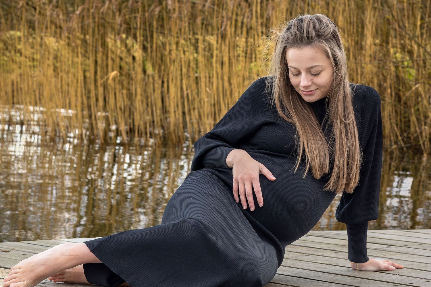 Foto af gravid kvinde i sort kjole. På træbro foran siv. Hun hviler hånden på maven. Et roligt, naturligt graviditetsportræt udendørs.