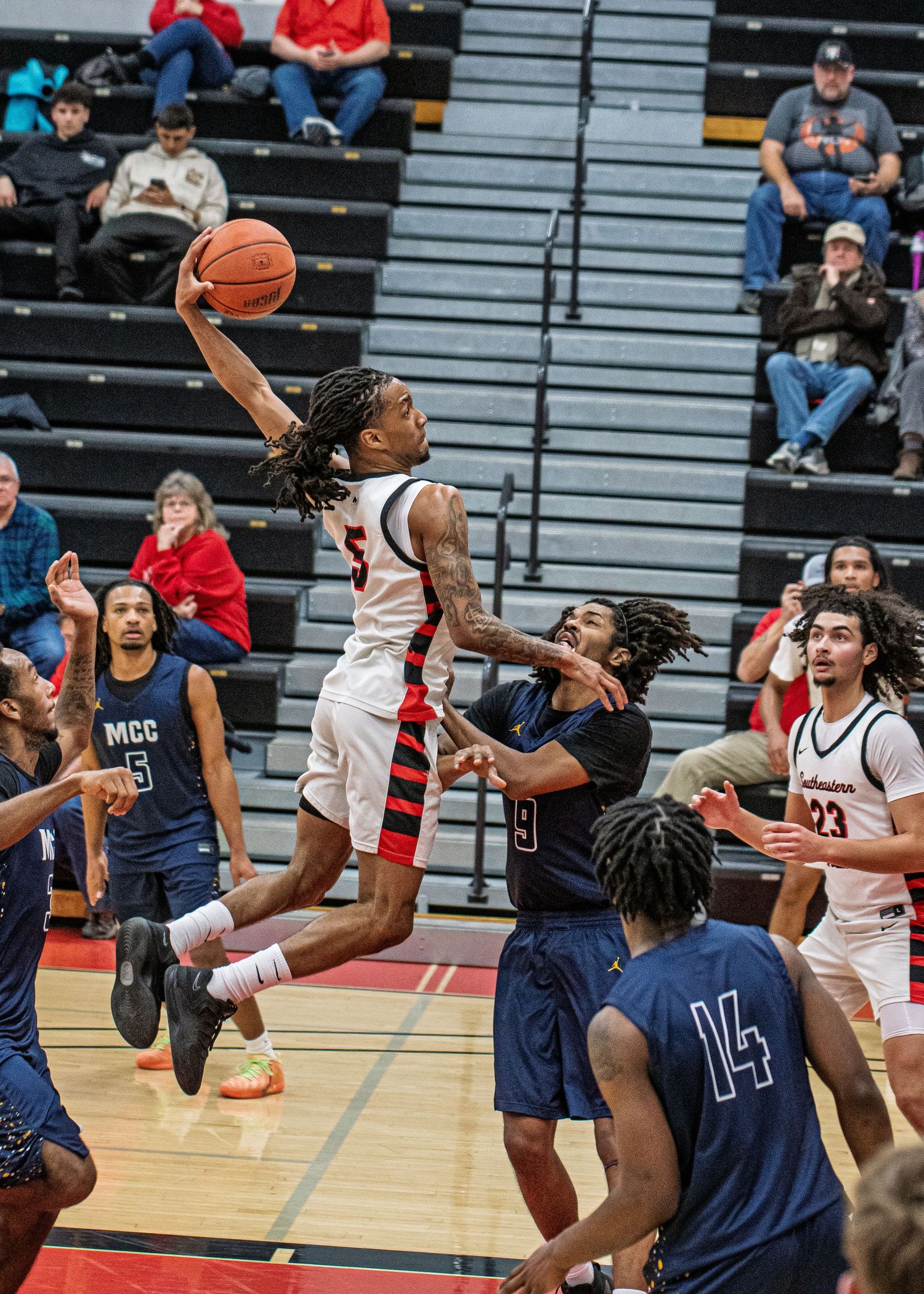 A basketball player in red leaps for a layup against MCC defenders in an indoor gymnasium game.