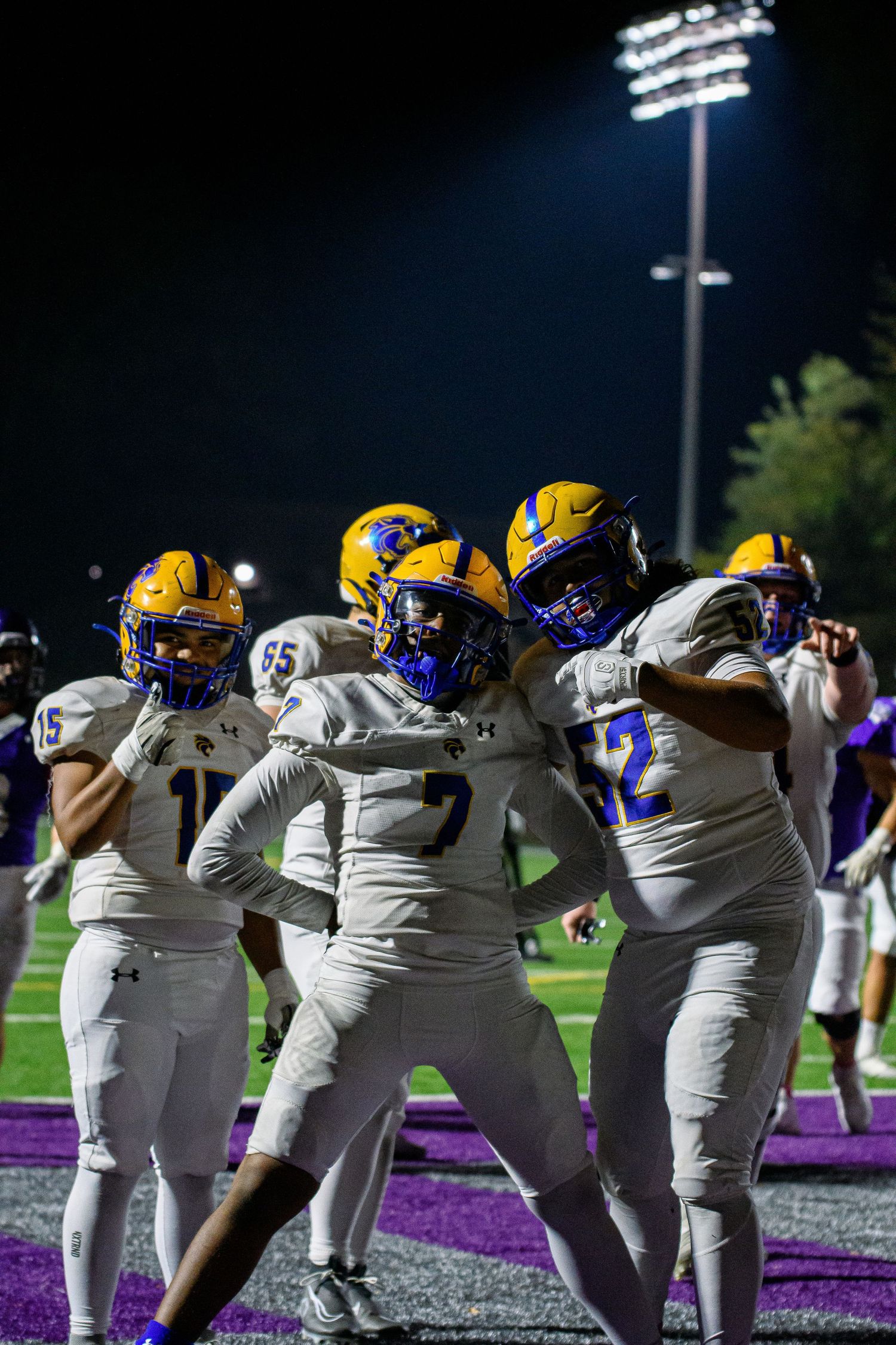 High school football players in gold helmets and white uniforms celebrate together under stadium lights at a night game.