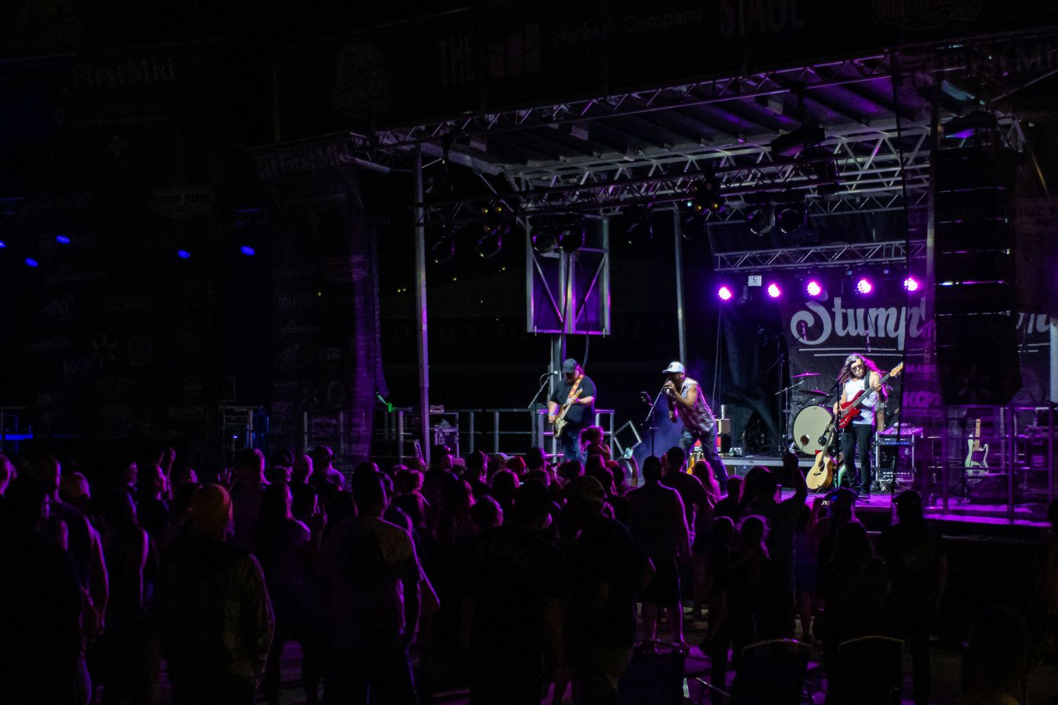 Live band performs on a lit stage before a large crowd at an outdoor Stump concert at night.