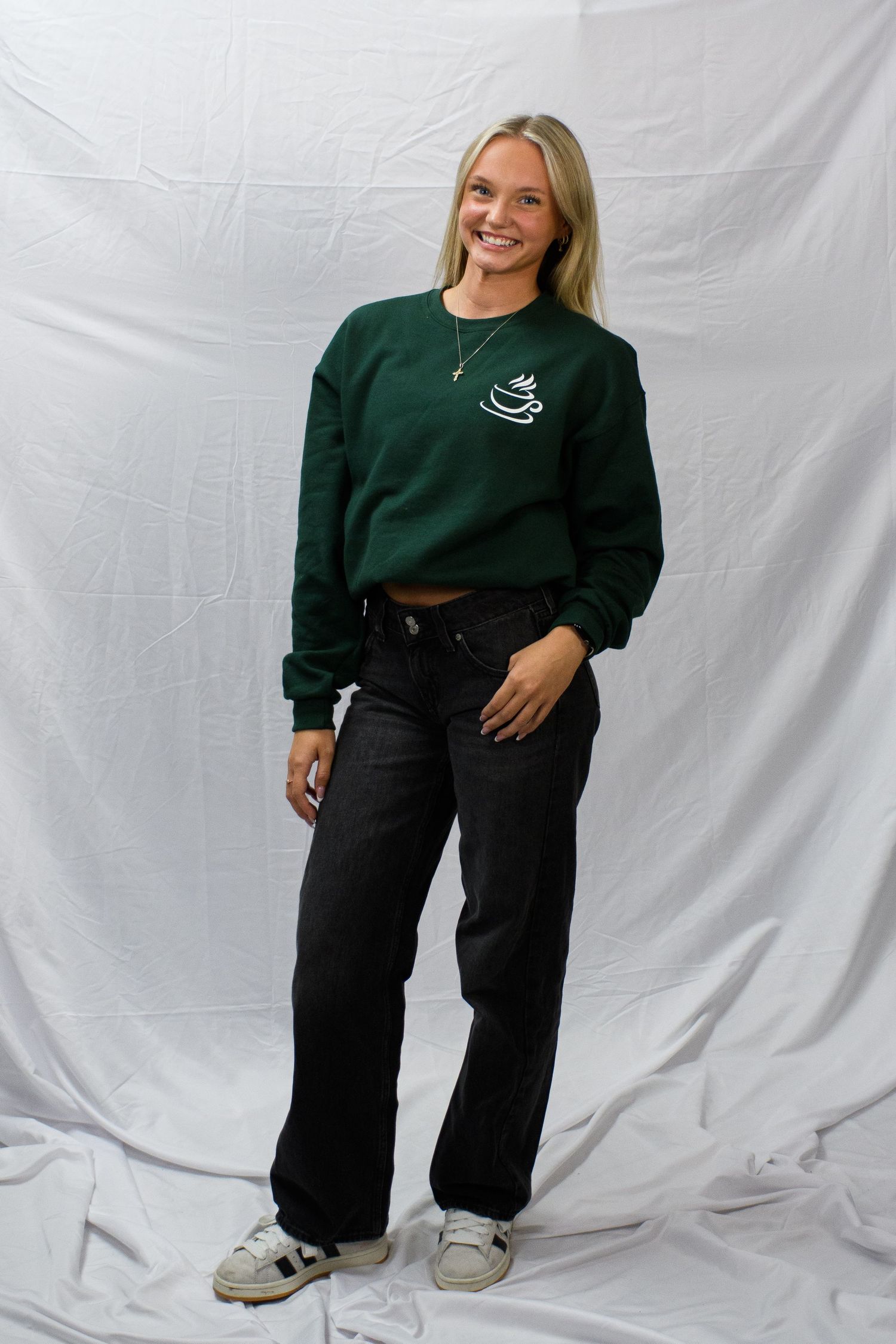 Woman smiling in green cropped sweatshirt with logo, black wide-leg jeans, and white sneakers against white backdrop.