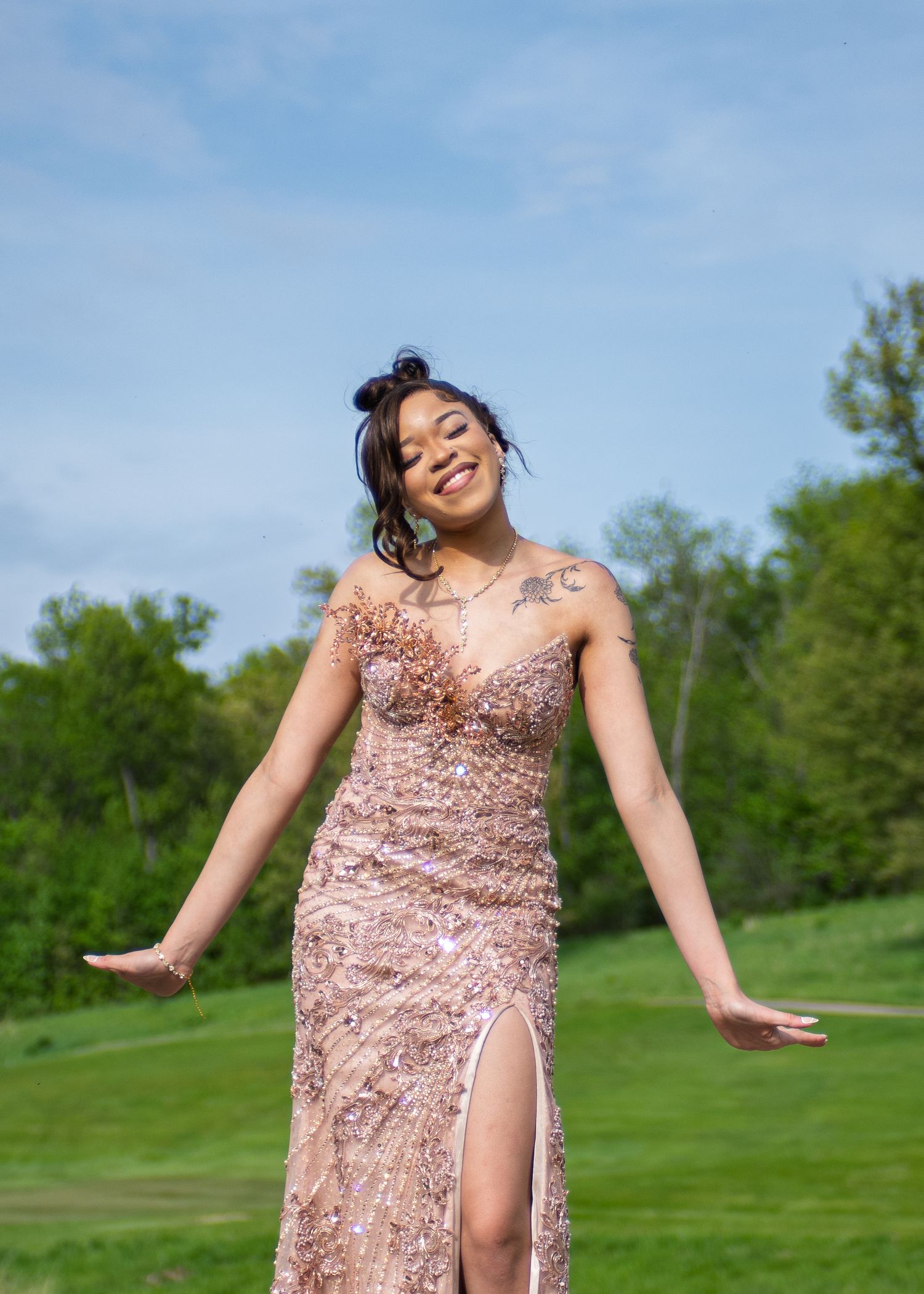 Young woman in a rose gold sequin gown poses outdoors on a green lawn under a blue sky.