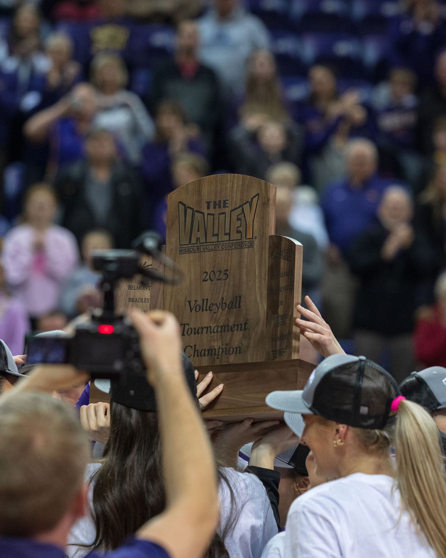 Players celebrate holding the 2025 Valley Volleyball Tournament Champion trophy aloft in a packed arena.