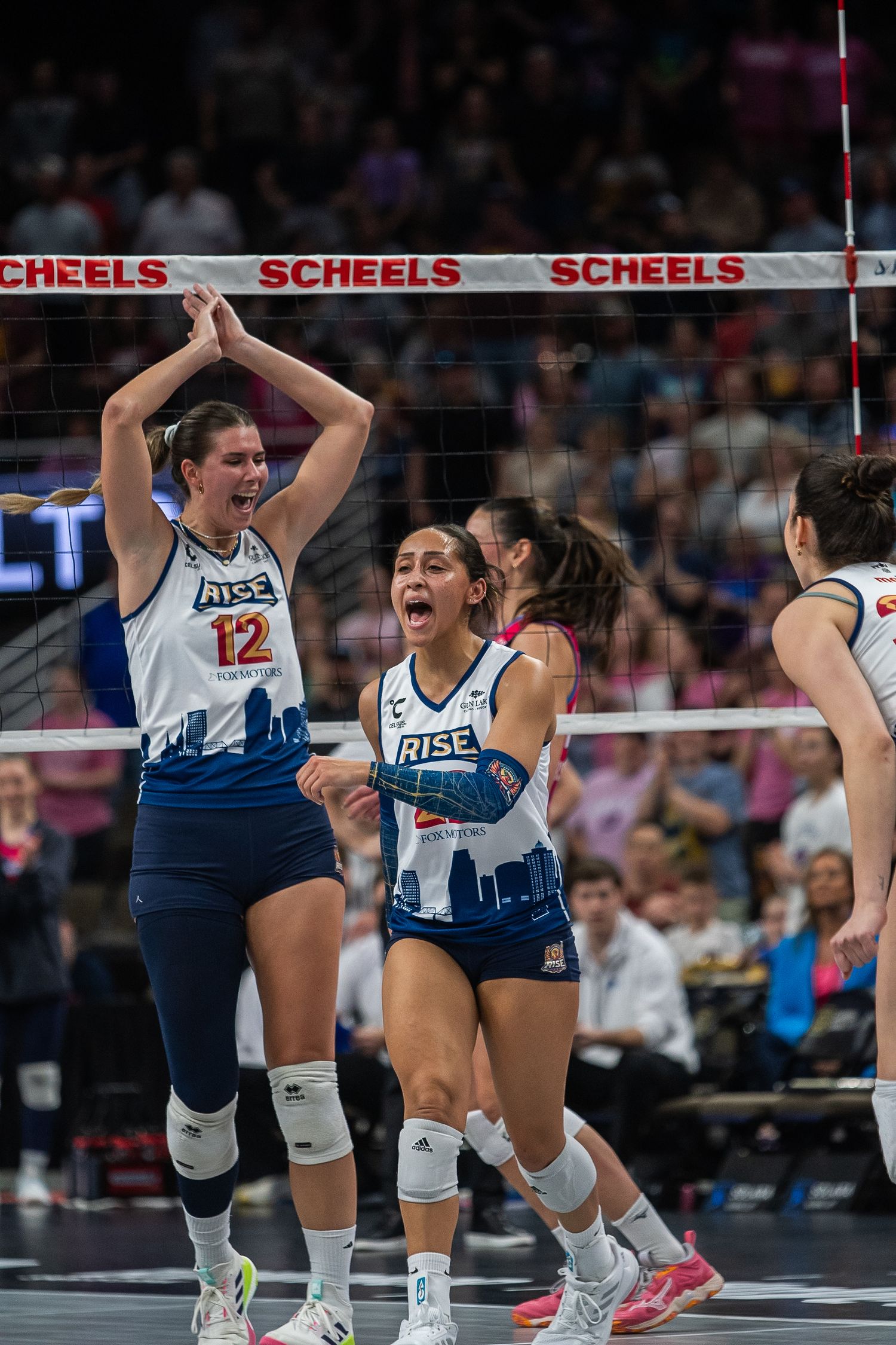 RISE volleyball players celebrate a point during a match, wearing white and blue uniforms with number 12 visible.