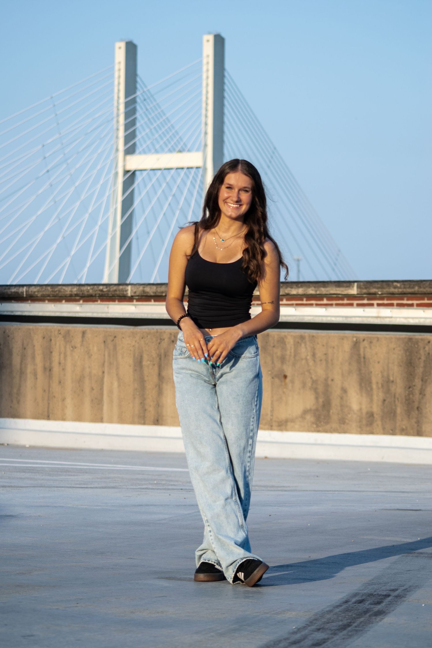 A person in a black tank top and light blue jeans poses casually against a concrete wall with a bridge in the background.