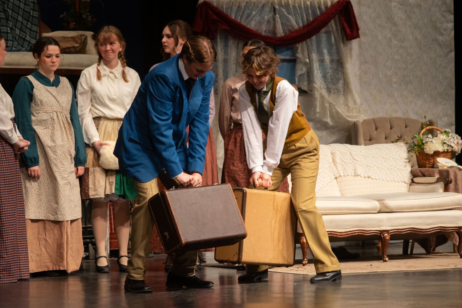 Actors perform a theatrical stage play scene with period costumes, carrying a suitcase on a decorated set.