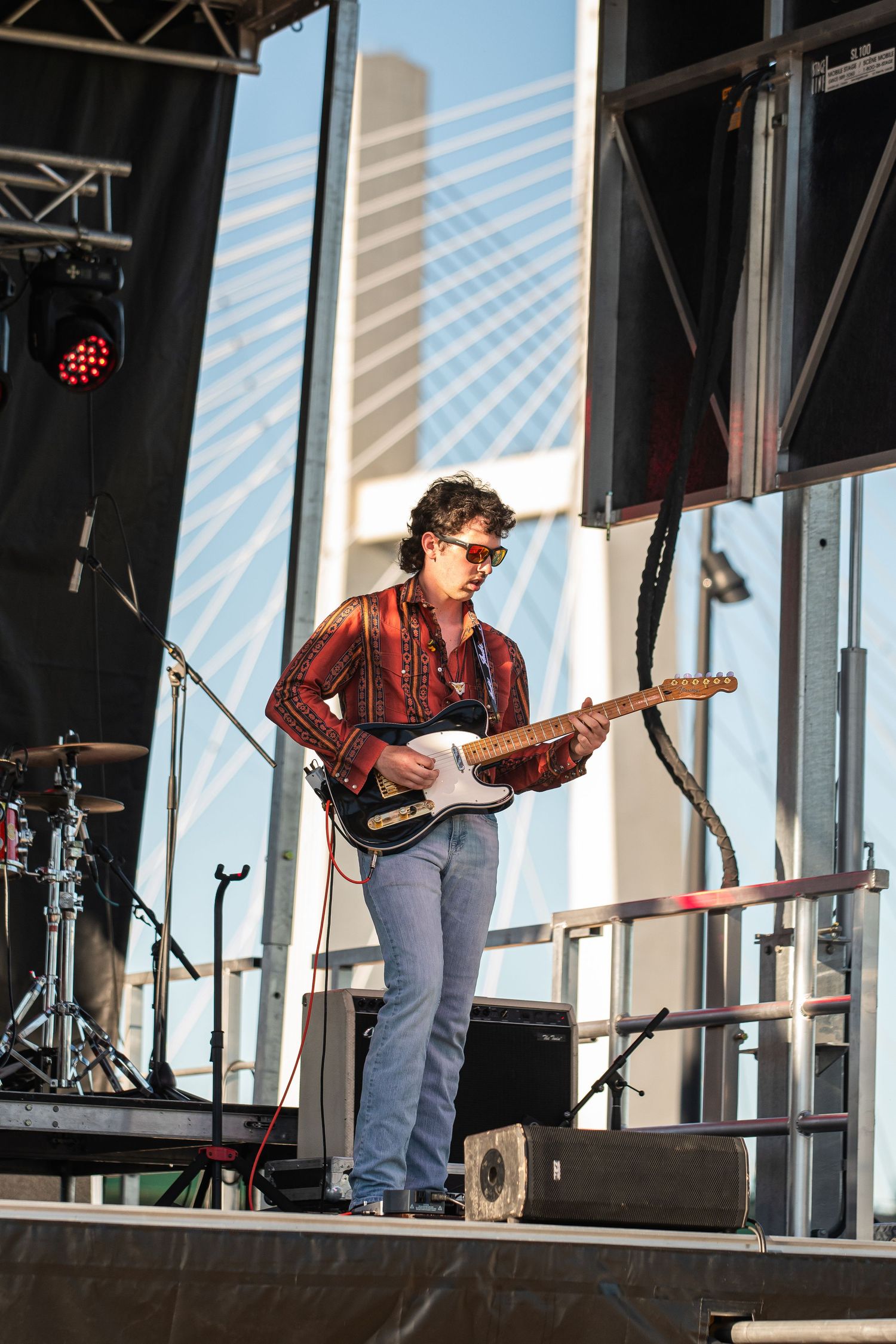 Young guitarist in plaid shirt and sunglasses performs on outdoor stage with bridge backdrop.