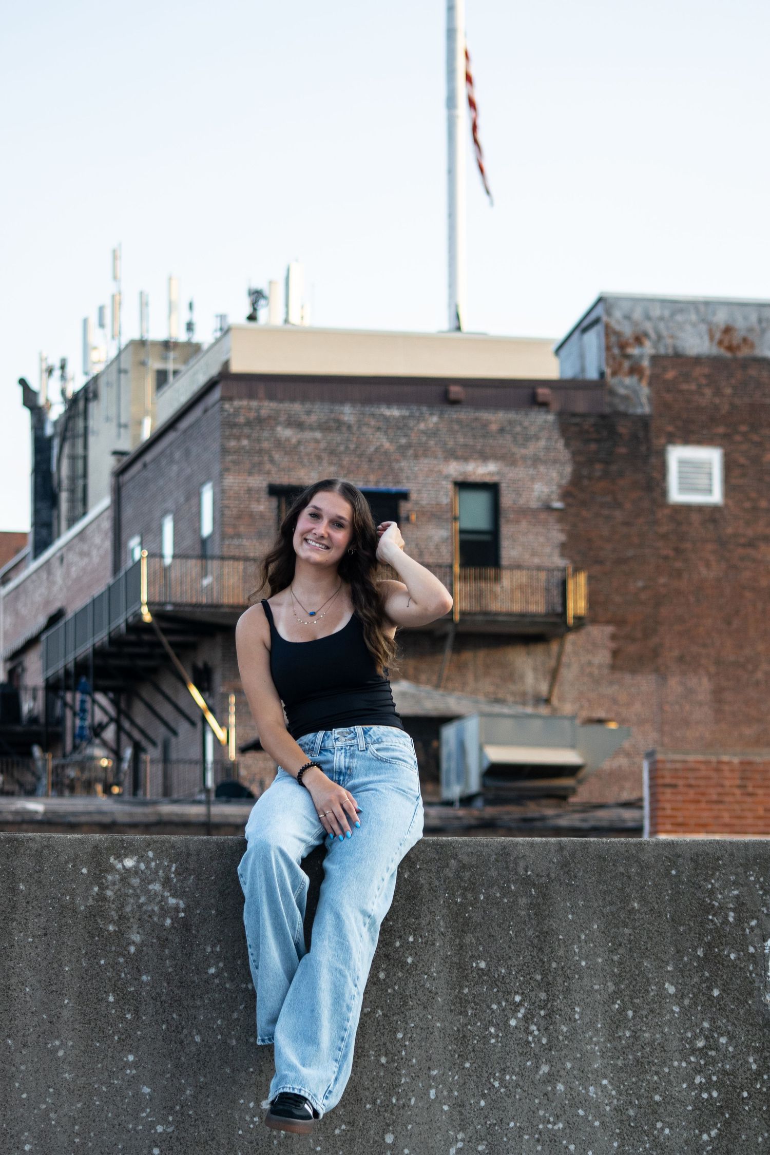 Young woman in black tank top and wide-leg jeans sits on a rooftop wall, smiling, with brick buildings and US flag behind her.