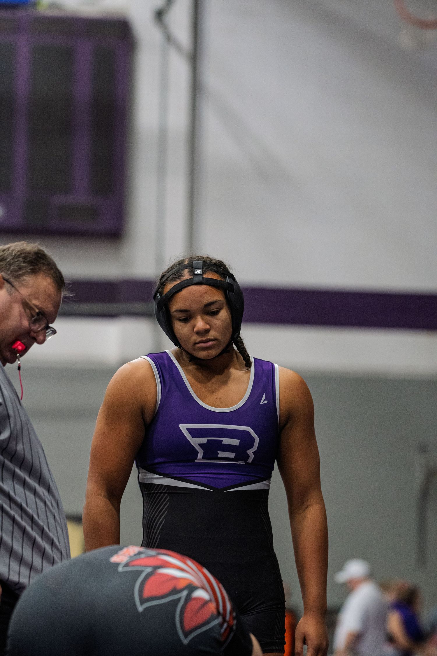 Female wrestler in purple singlet with headgear prepares for match at indoor wrestling tournament.