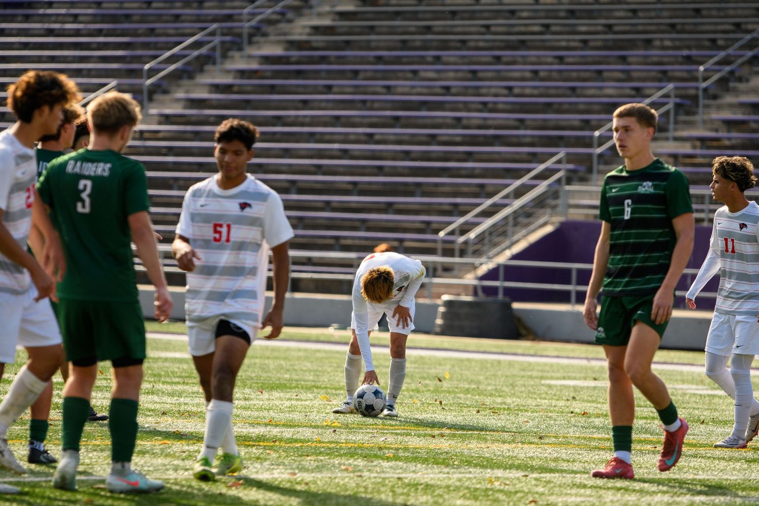 Soccer players compete for the ball during a match on a sunny day at a stadium field.
