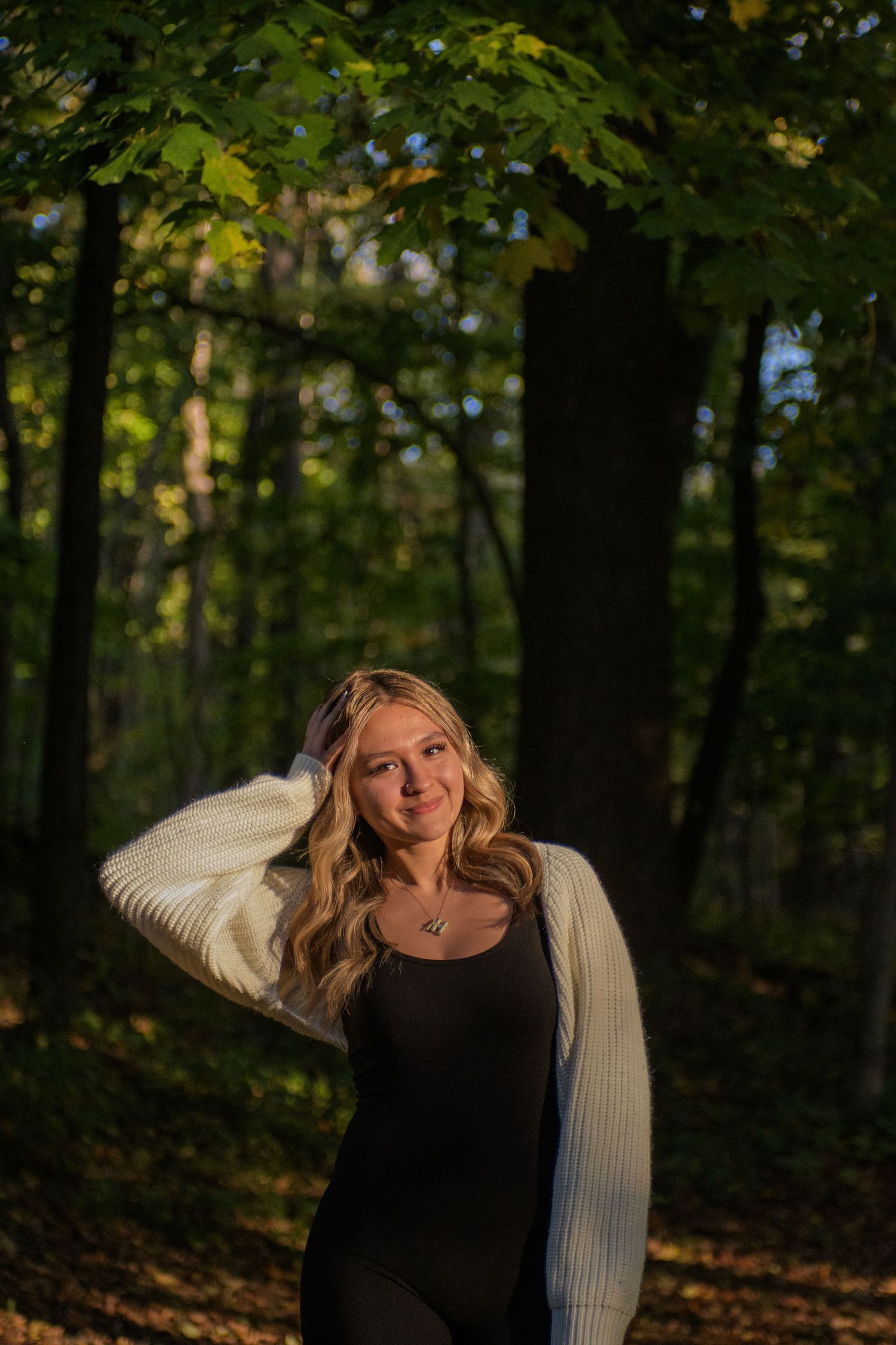 Young blonde woman in white cardigan and black outfit posing in sunlit autumn forest.