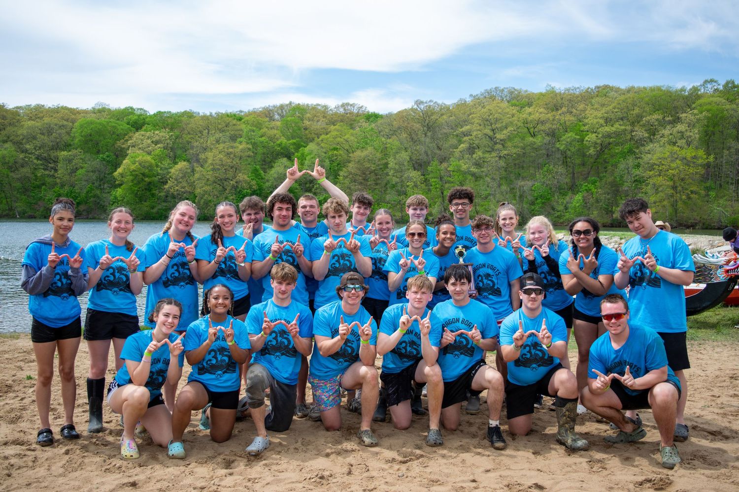 A large group in matching blue t-shirts poses together outdoors on a sunny day with trees in the background.