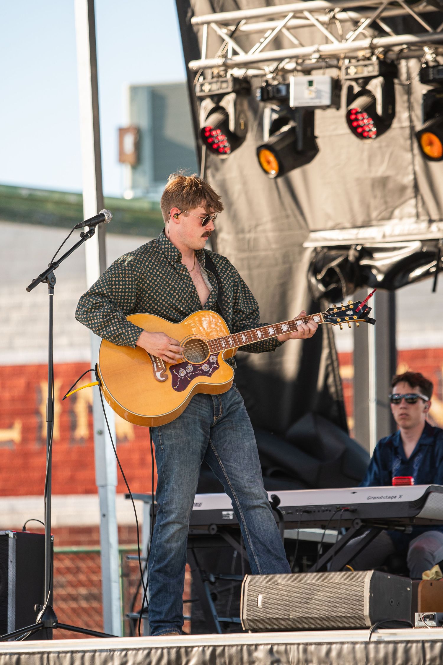 Male guitarist performing on outdoor stage with acoustic guitar, microphone, and stage lights at a live music concert.