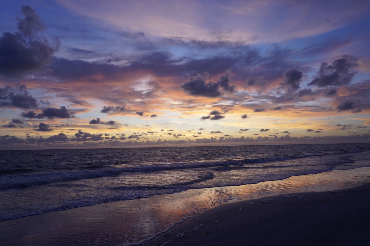 Stunning beach sunset with purple and orange clouds reflecting on wet sand and rolling waves.