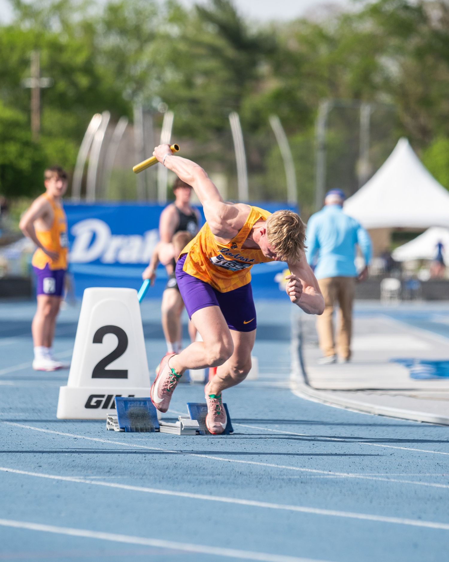Athlete in purple and gold uniform bursting from starting block 2, holding relay baton at outdoor track meet.