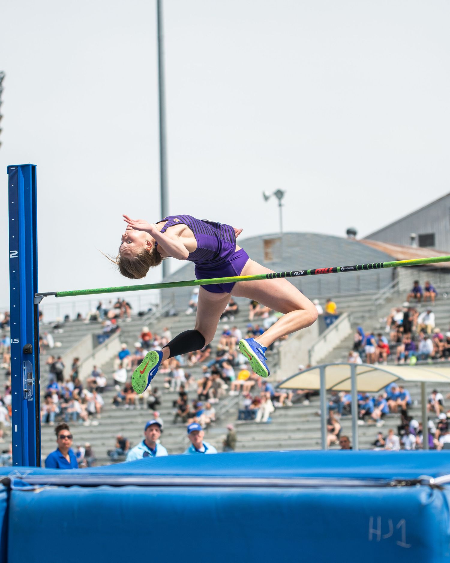 Athlete in purple uniform performs high jump, clearing green bar at outdoor track meet with spectators in background.