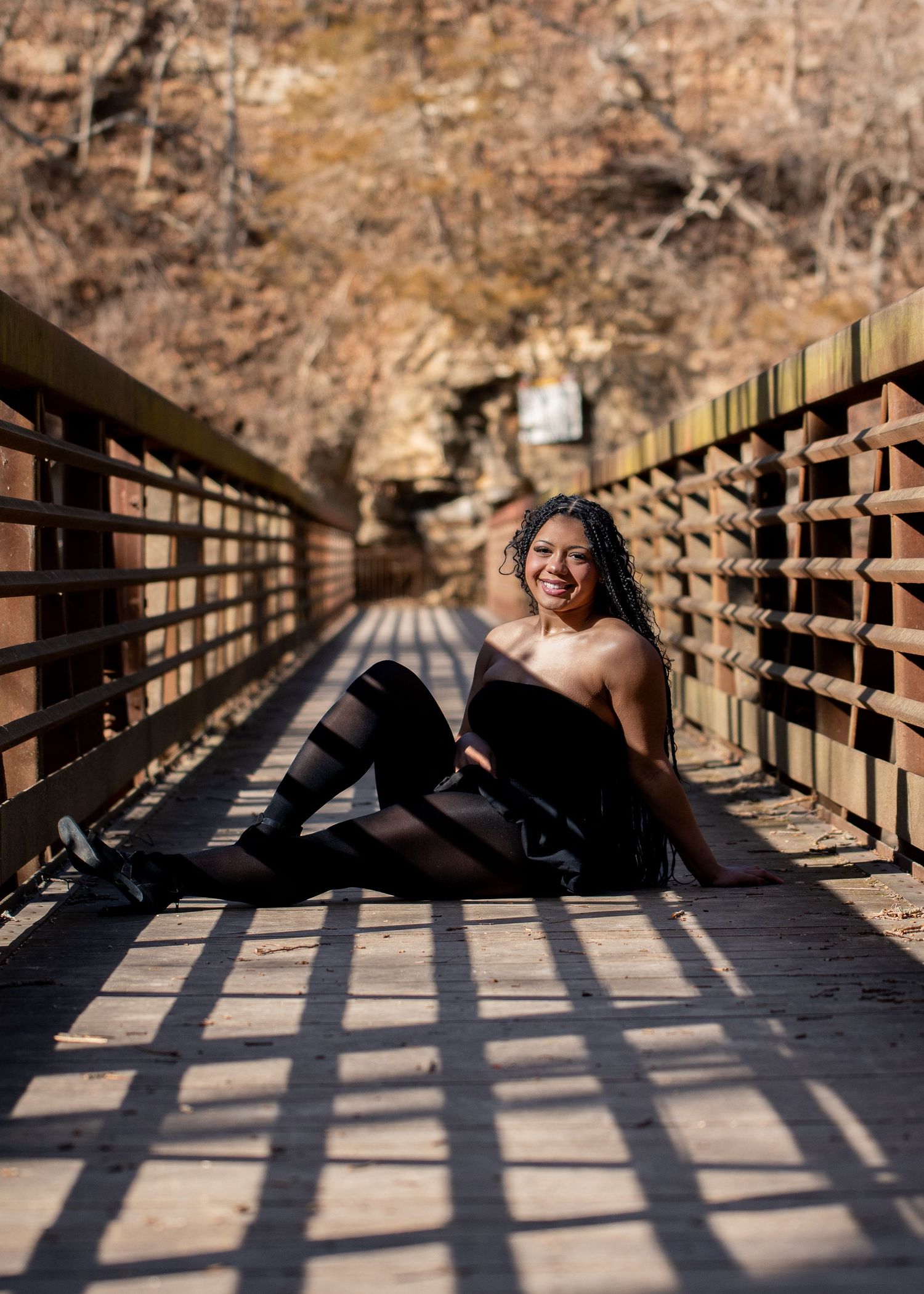 Woman in black outfit sitting on a wooden bridge surrounded by autumn foliage, smiling joyfully.
