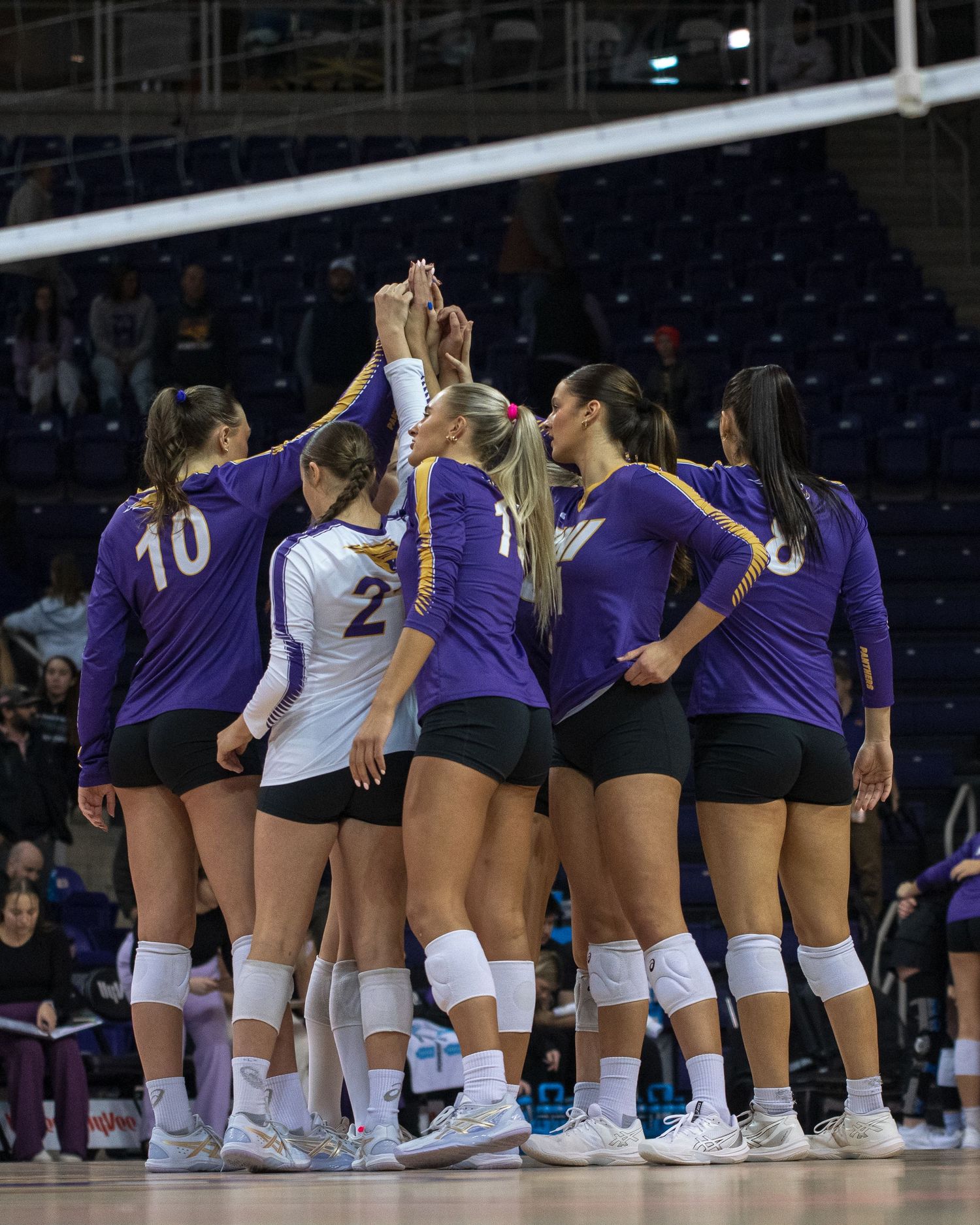 A volleyball team huddles together in a circle wearing purple and white uniforms during a game.