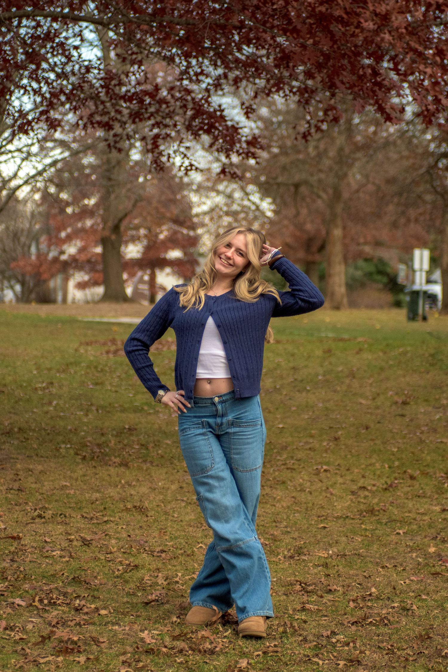 Young blonde woman in blue cropped sweater and wide-leg jeans posing outdoors in a park with autumn trees.