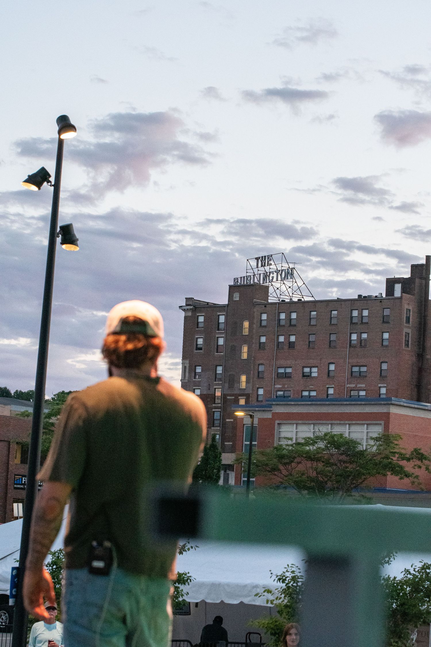 Man in cap stands facing The Burlington Hotel at dusk, with street lamps and cloudy sky in background.