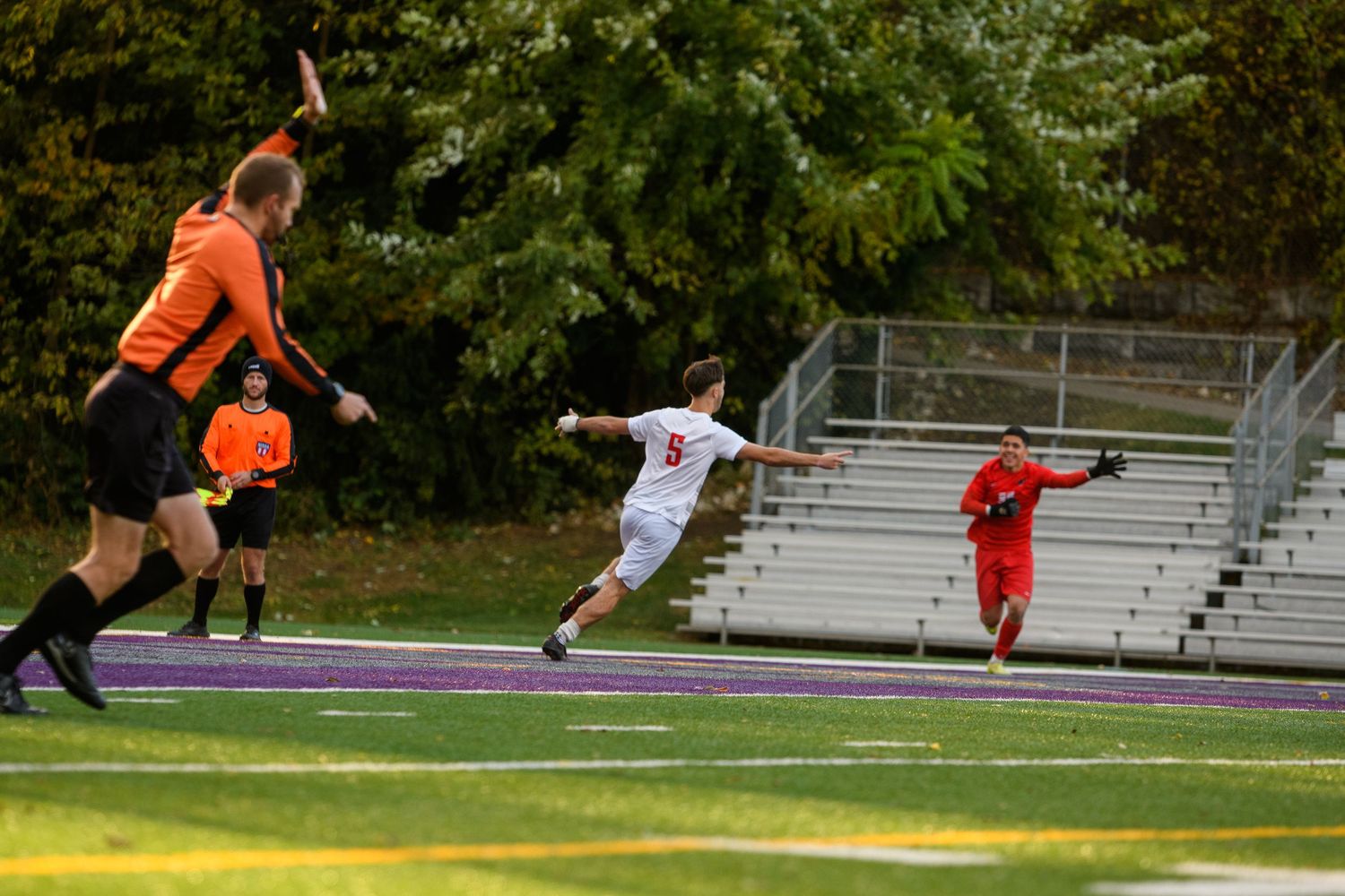 Soccer players compete on a purple turf field during a youth match, with a referee raising their hand nearby.
